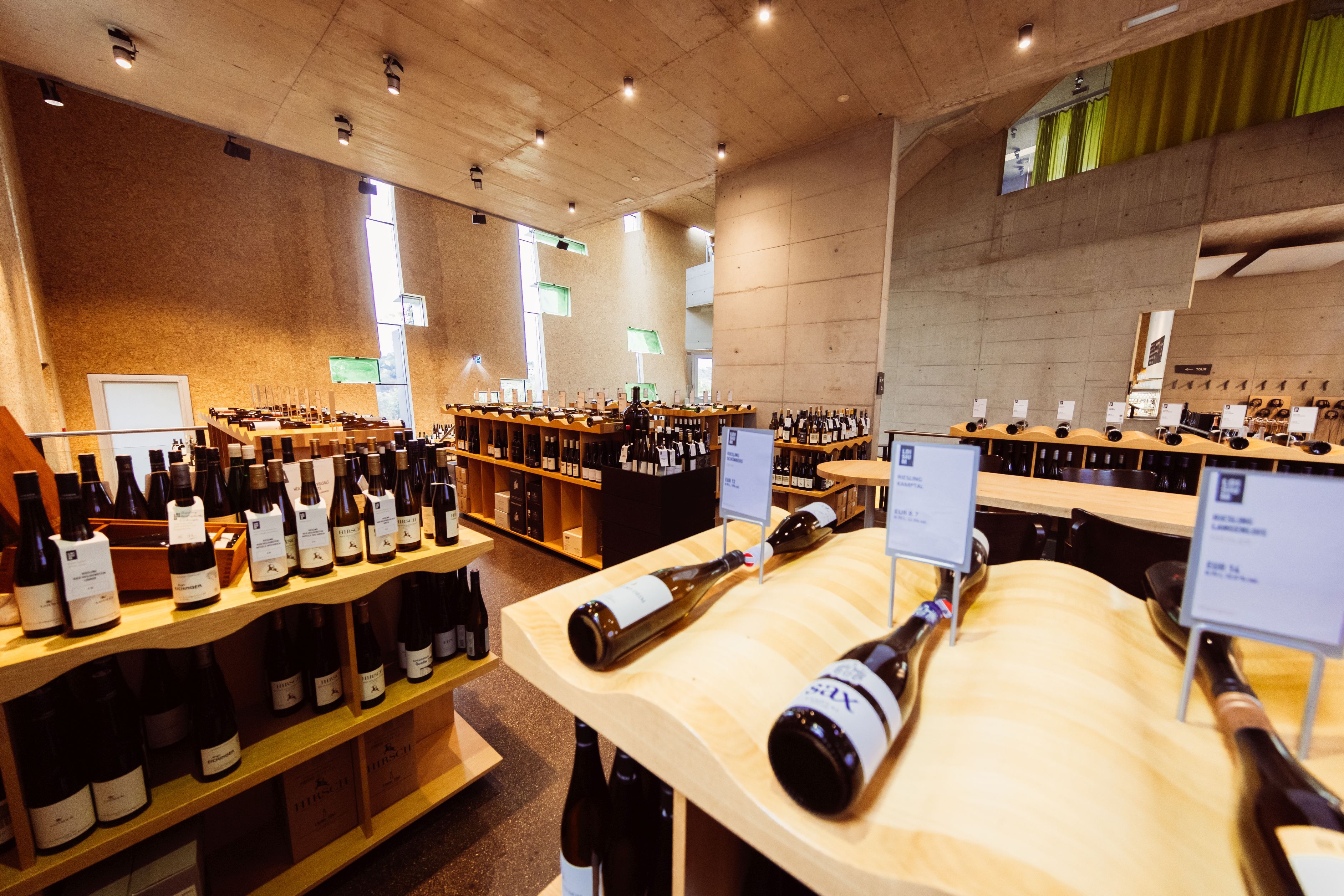 Innenansicht der Vinothek Steininger Weinwelt mit zahlreichen Weinflaschen auf Holzregalen und Tischen in modernem Betonambiente.