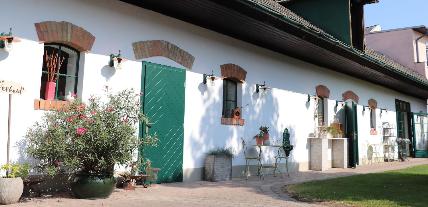 Ein weißes Gebäude mit grünen Türen und Fenstern, umgeben von Pflanzen und Gartenmöbeln.