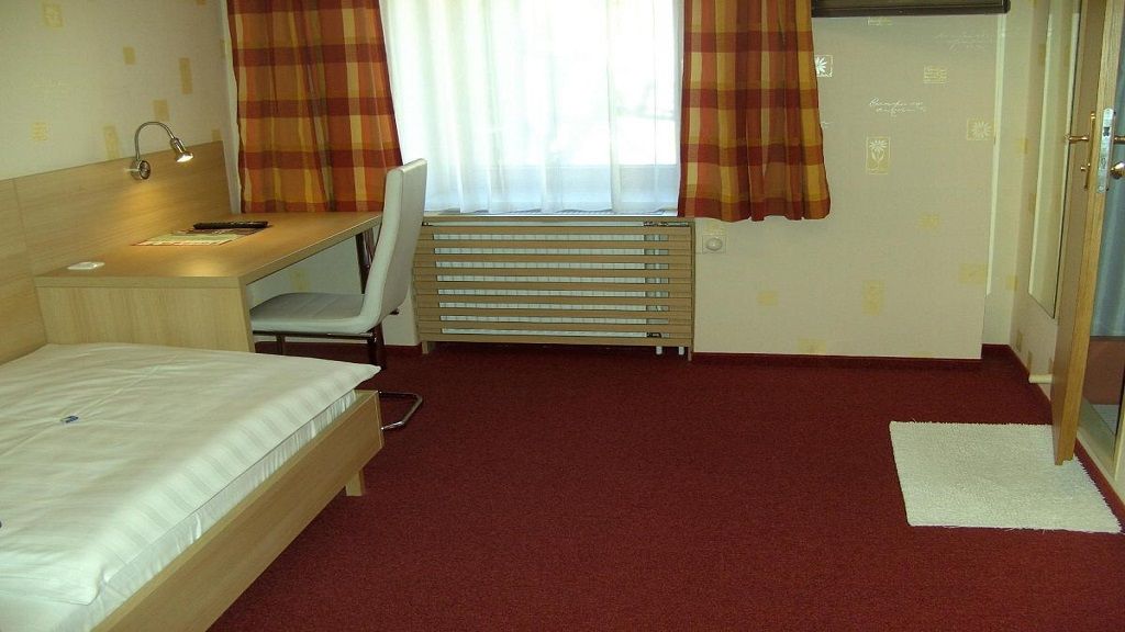 Ein einfach eingerichtetes Hotelzimmer mit Einzelbett, Schreibtisch und Stuhl. Rote Teppichboden und karierte Vorhänge.