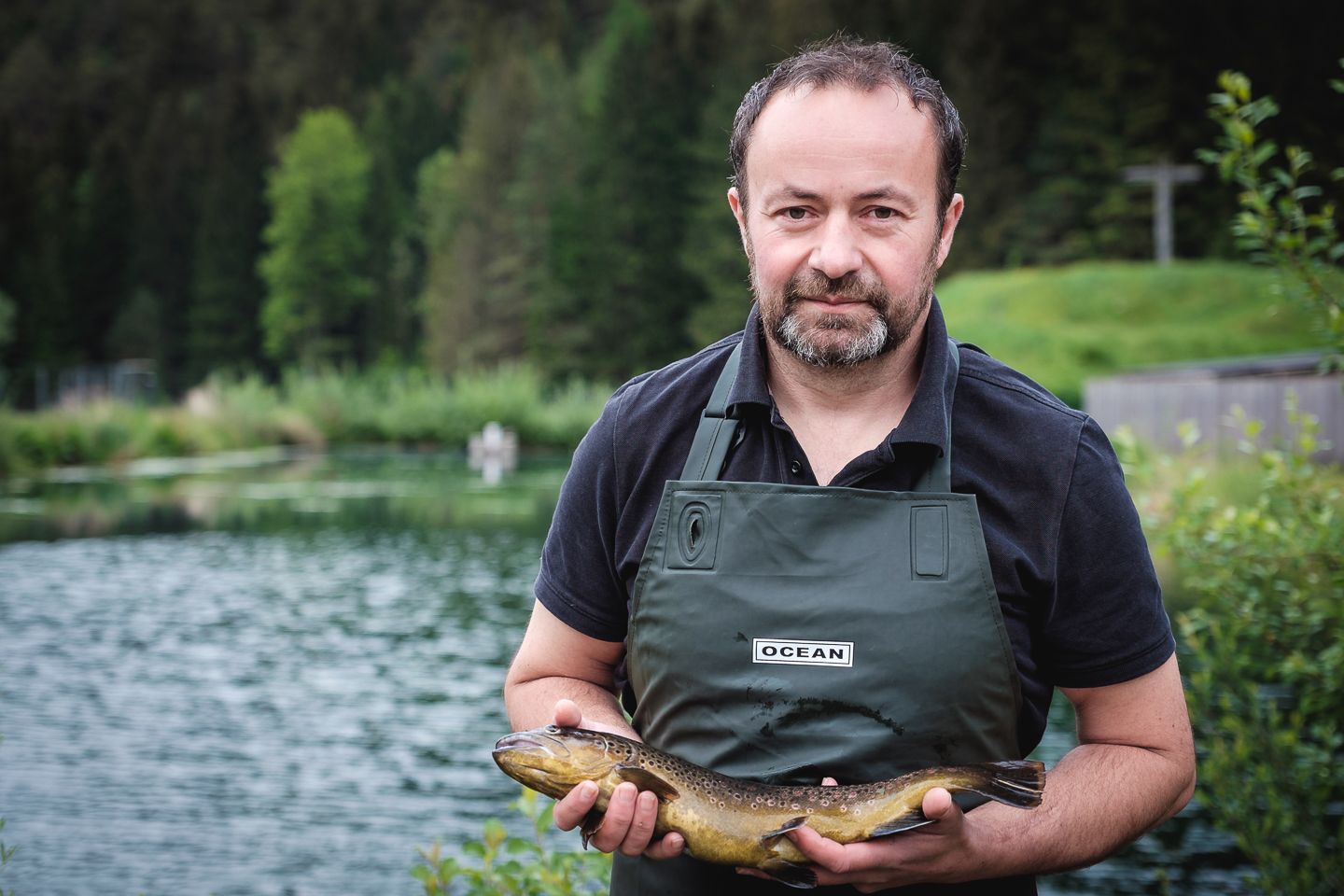 Ein Mann in grüner Schürze hält einen Fisch vor einem Teich.