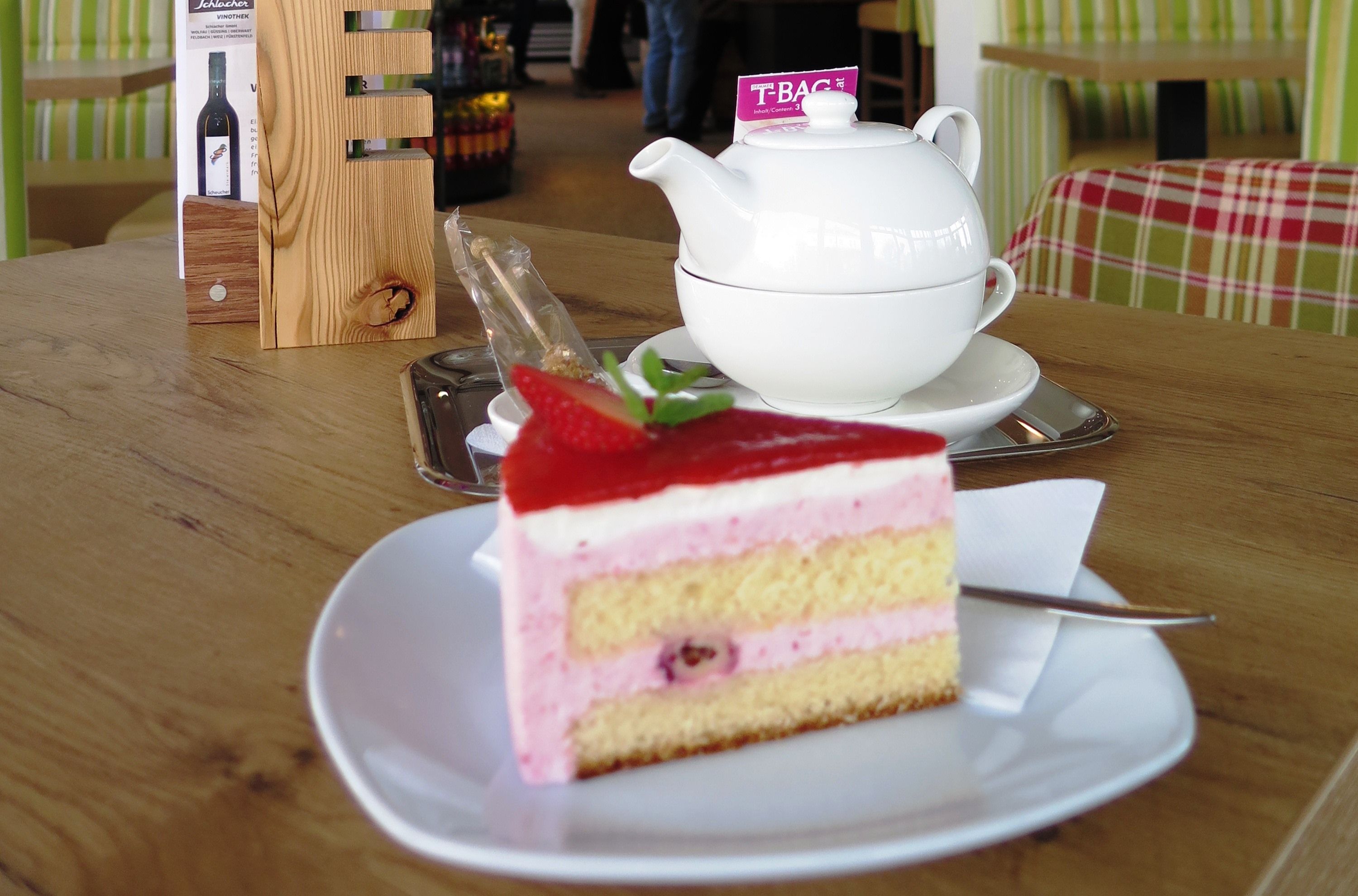Ein Stück Erdbeerkuchen auf einem Teller mit einer Teekanne im Hintergrund auf einem Holztisch.