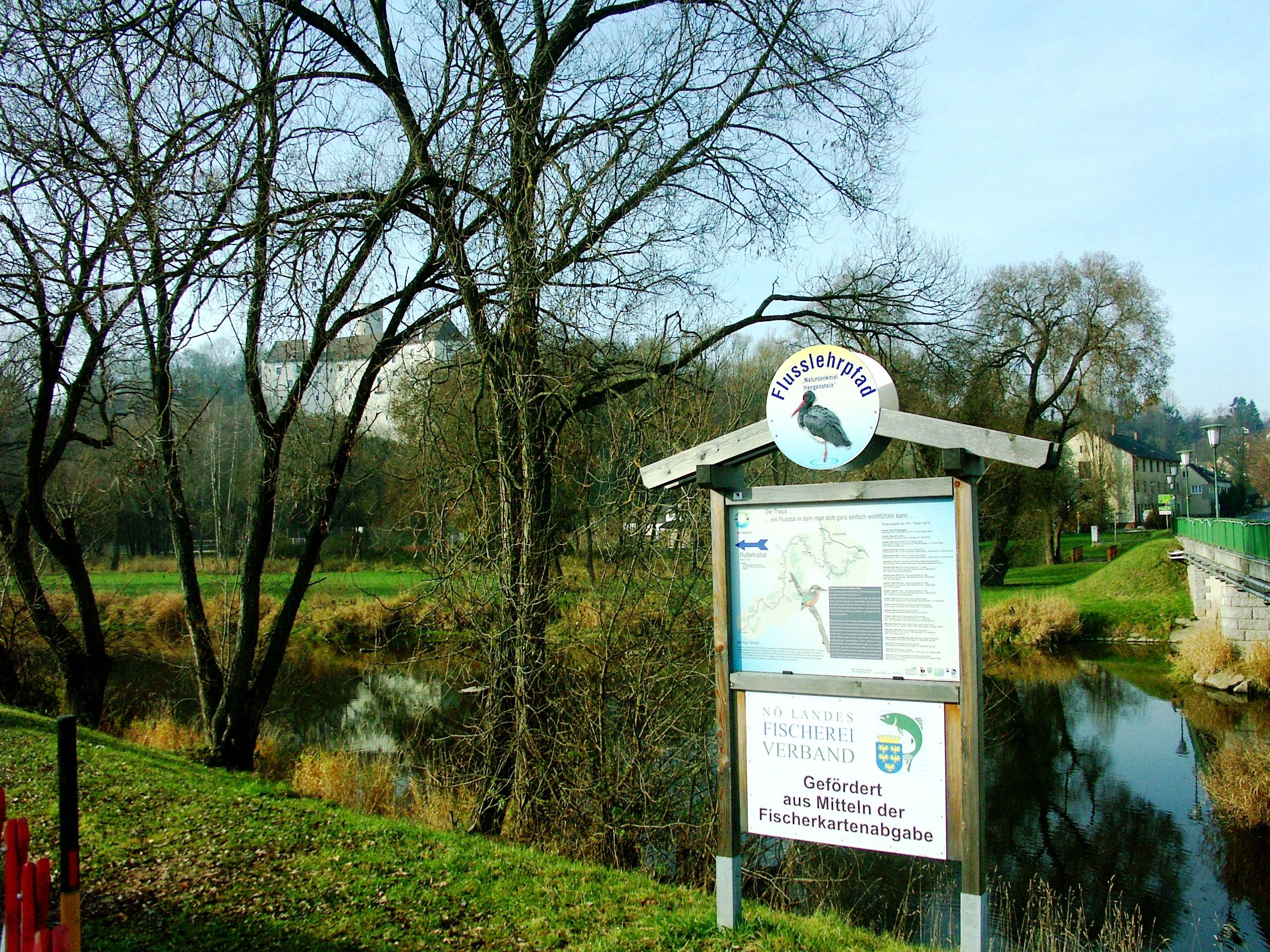 Ein Flusslehrpfad-Schild an einem Flussufer mit kahlen Bäumen im Hintergrund.