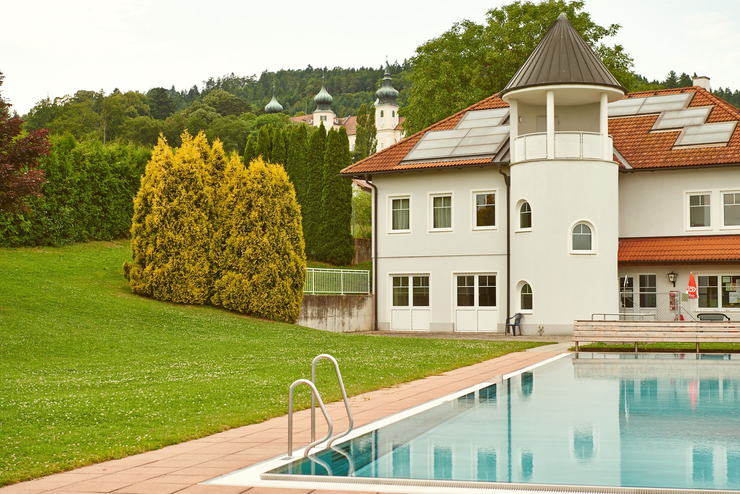 Außenansicht einer Freizeitanlage mit Pool und Gebäude, im Hintergrund Bäume und Kirchtürme.