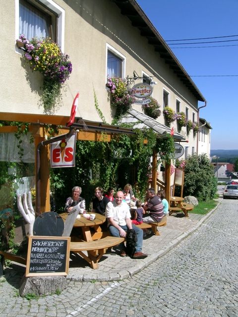 Eine Gruppe von Menschen sitzt vor einem Gasthof an Holztischen auf einer gepflasterten Straße.