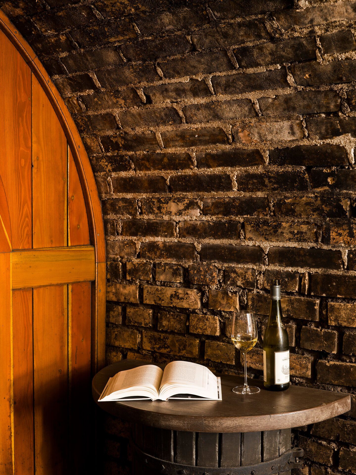 In einem gemütlichen Weinkeller, umgeben von rustikalen Ziegelwänden, lädt ein Glas Weißwein dazu ein, die Seele baumeln zu lassen. Ein aufgeschlagenes Buch verspricht entspannende Lesestunden, während die Atmosphäre des Ortes die Sinne verwöhnt. Hier wird die Kultur des Weins auf besondere Weise erlebbar.