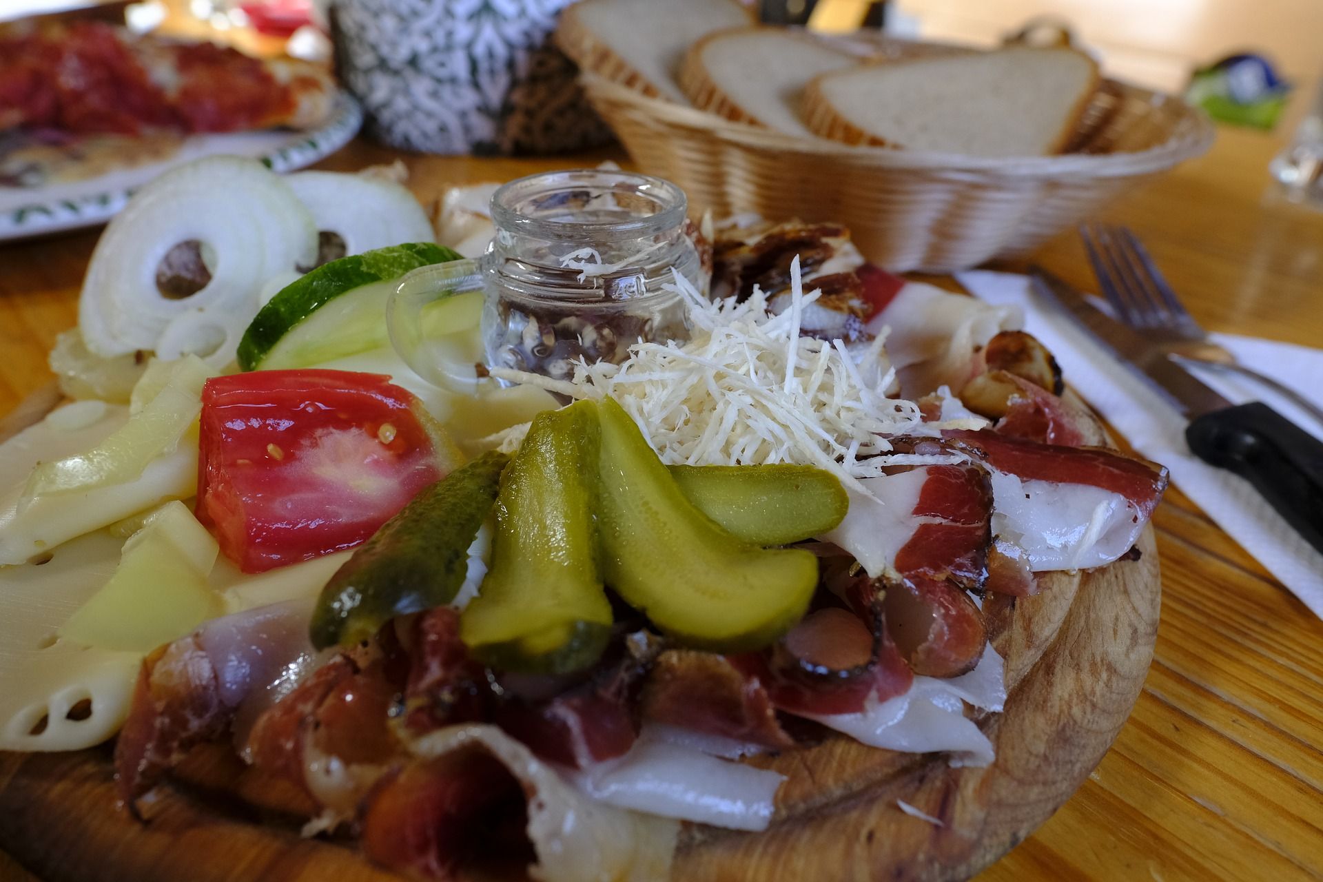 Ein rustikales Brett mit Käse, Schinken, Gurken, Tomaten und Zwiebeln, serviert mit Brot im Hintergrund.