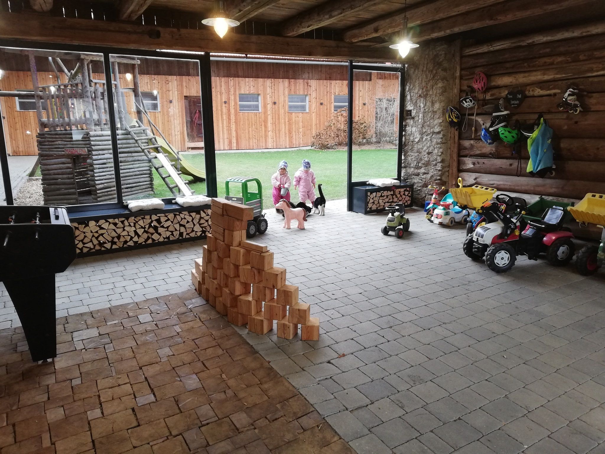 Innenansicht einer Spielescheune mit Spielzeugautos, Holzklötzen und zwei Kindern in Winterkleidung.