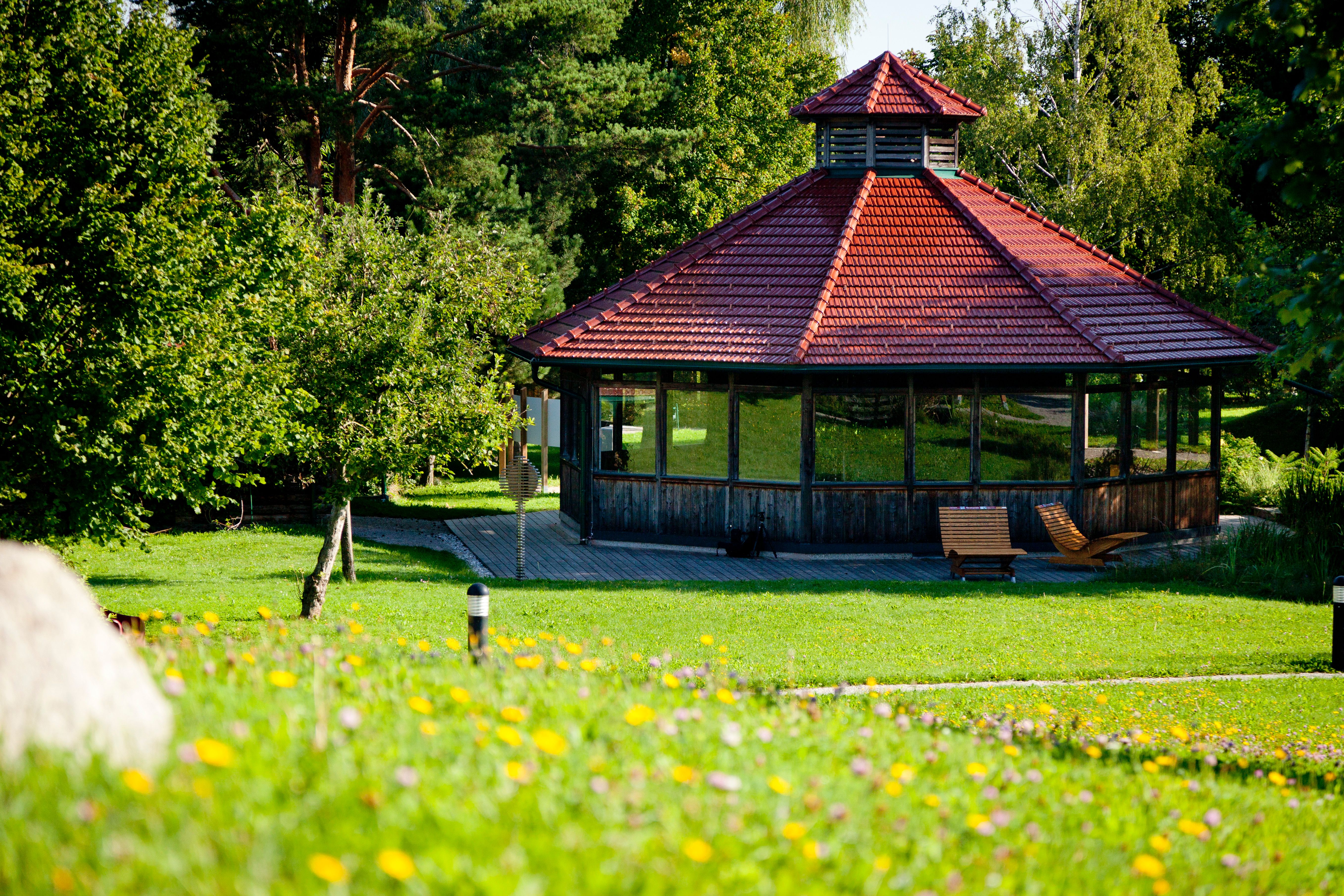Ein Pavillon mit rotem Dach in einem grünen Park, umgeben von Bäumen und Blumen.