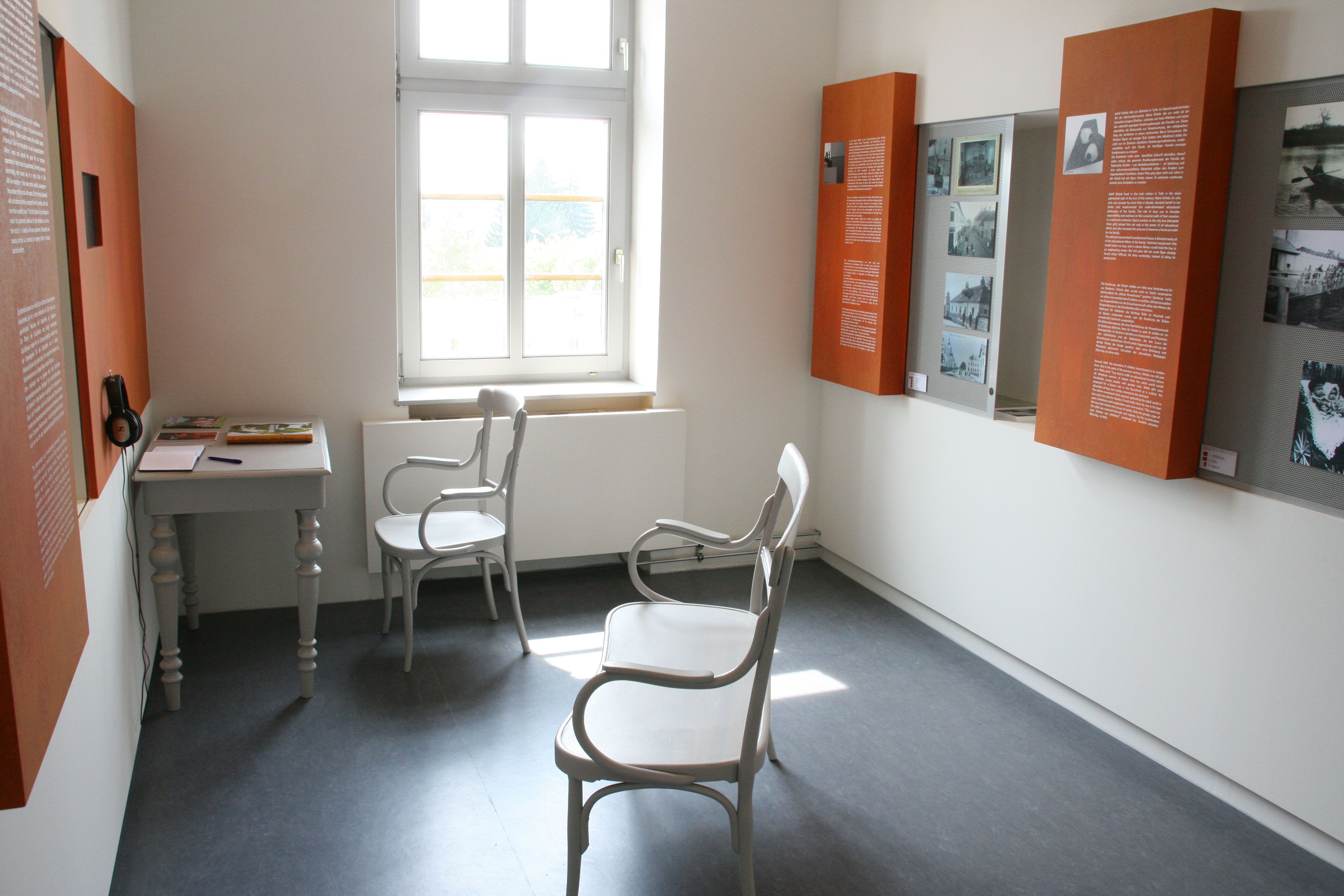 Innenansicht eines Museumsraums mit weißen Stühlen, einem Tisch und Infotafeln an den Wänden.
