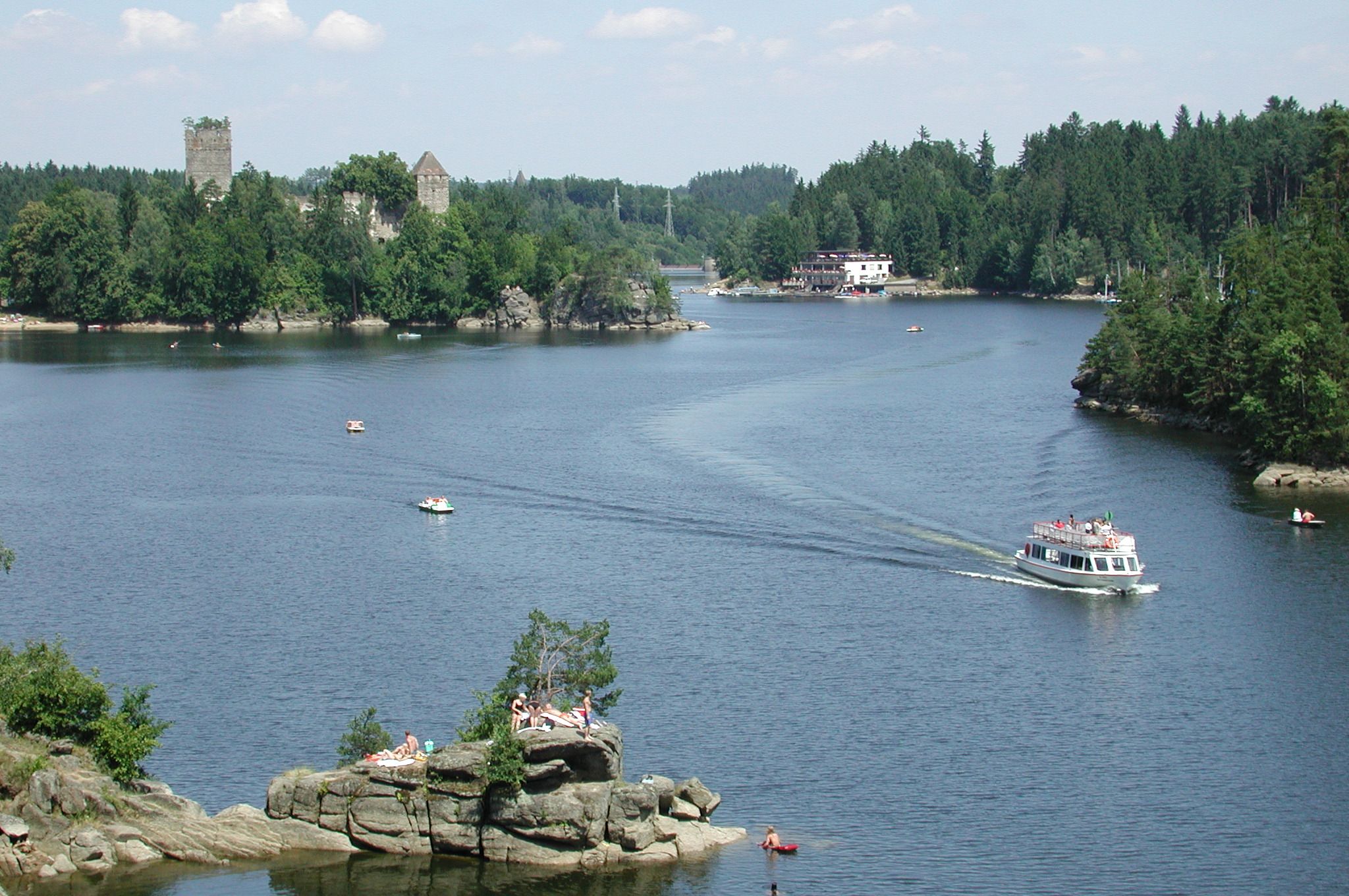 Ein Boot fährt auf einem See mit bewaldeten Ufern und einer Burg im Hintergrund.