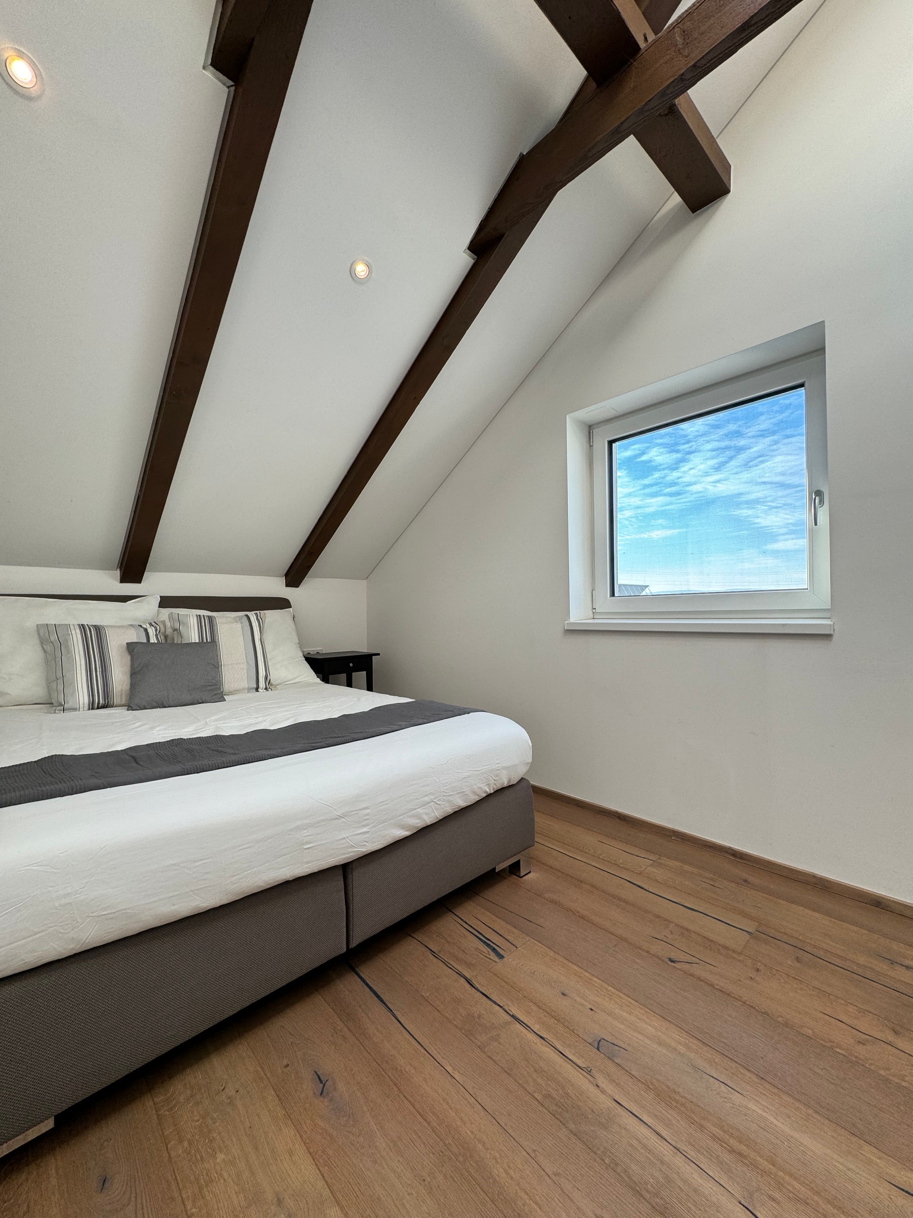 Modernes Schlafzimmer mit Holzbalkendecke, großem Bett und Fenster mit Blick auf blauen Himmel.
