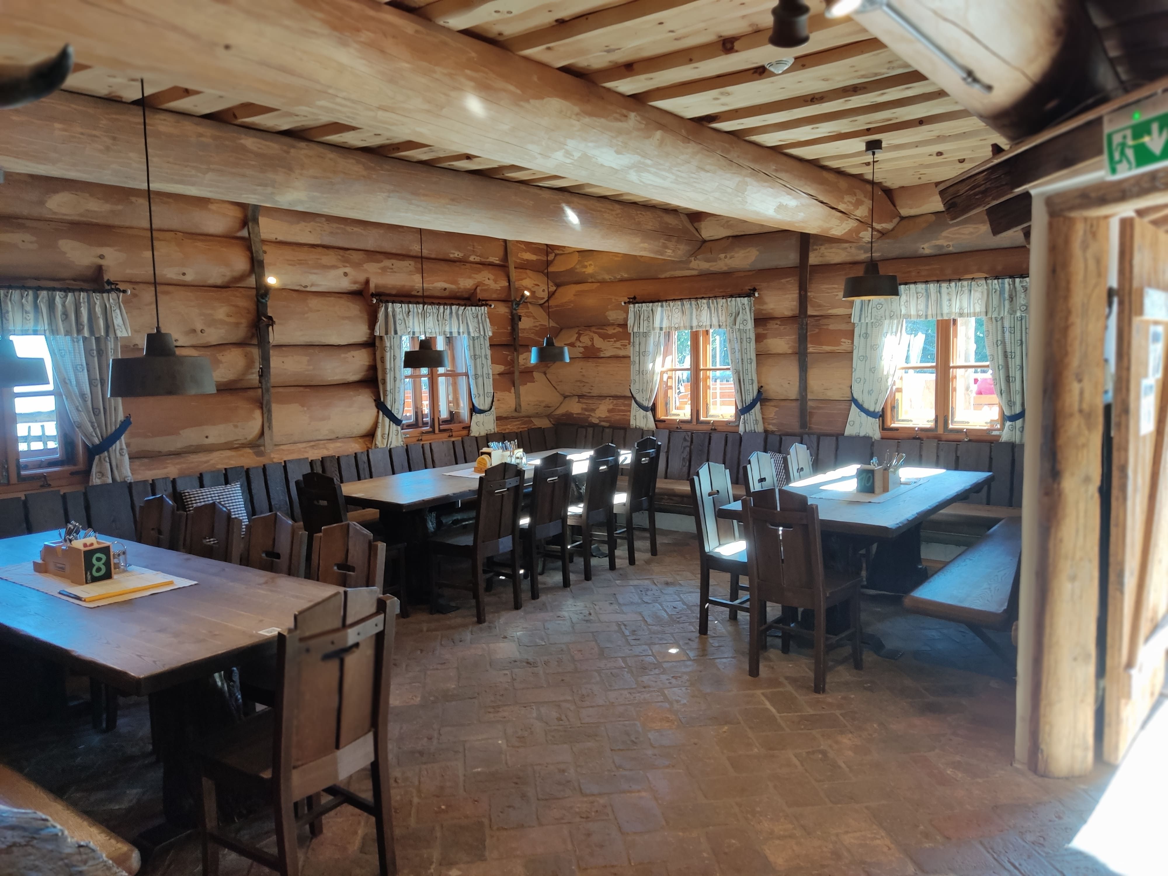 Innenansicht einer rustikalen Holzhütte mit Holztischen und Stühlen, dekoriert mit Vorhängen an den Fenstern.