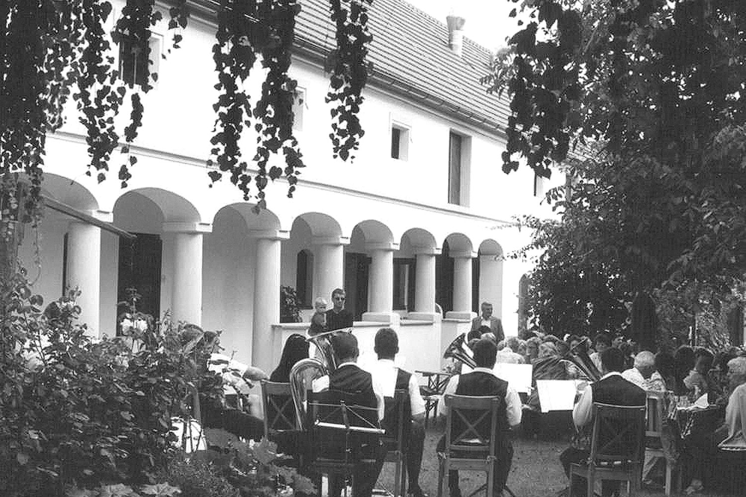 Eine Gruppe von Menschen sitzt im Freien vor einem Gebäude mit Säulen, während eine Blaskapelle spielt.