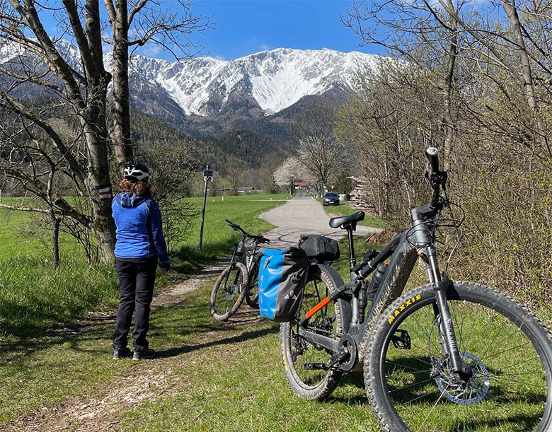 Eine Person mit Helm steht neben Fahrrädern auf einem Weg mit schneebedeckten Bergen im Hintergrund.