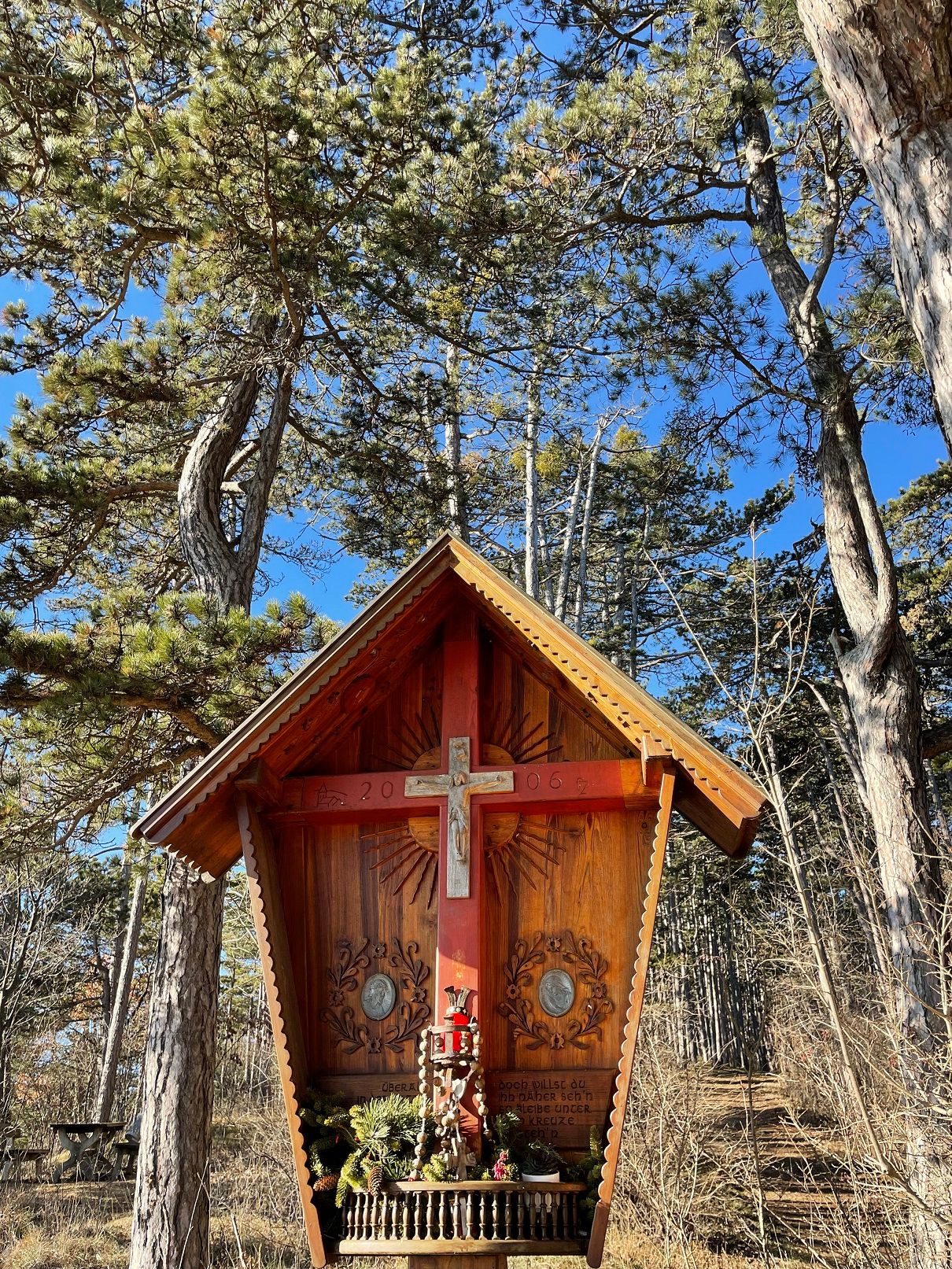 Holzkreuz mit Dach in einem Wald, umgeben von Bäumen und blauem Himmel.