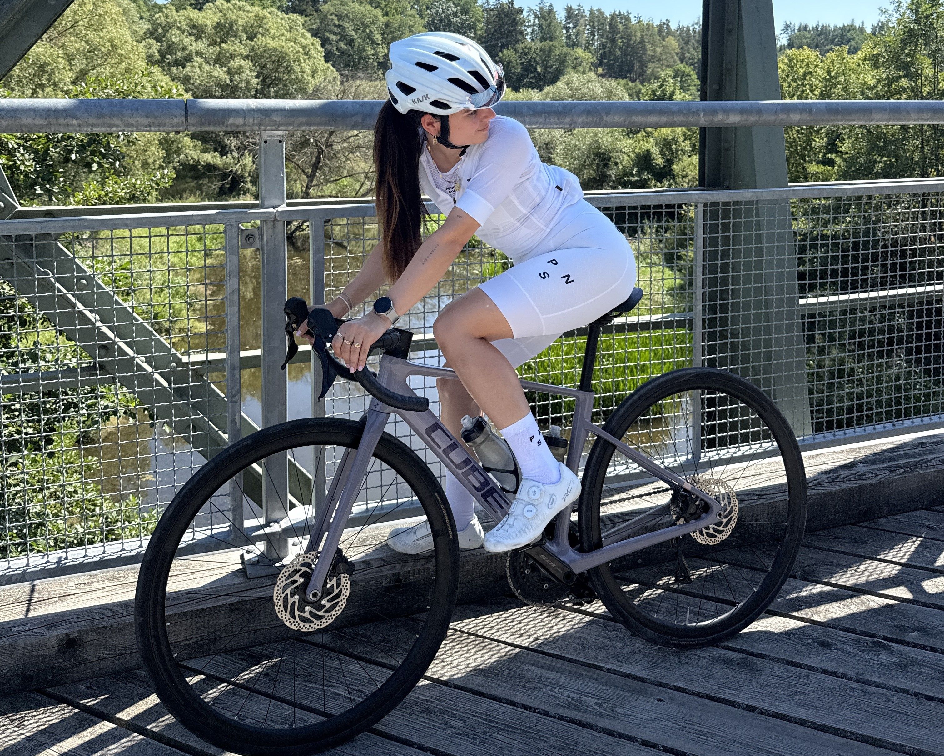 Frau in weißem Fahrradoutfit auf einer Brücke mit einem grauen Fahrrad.
