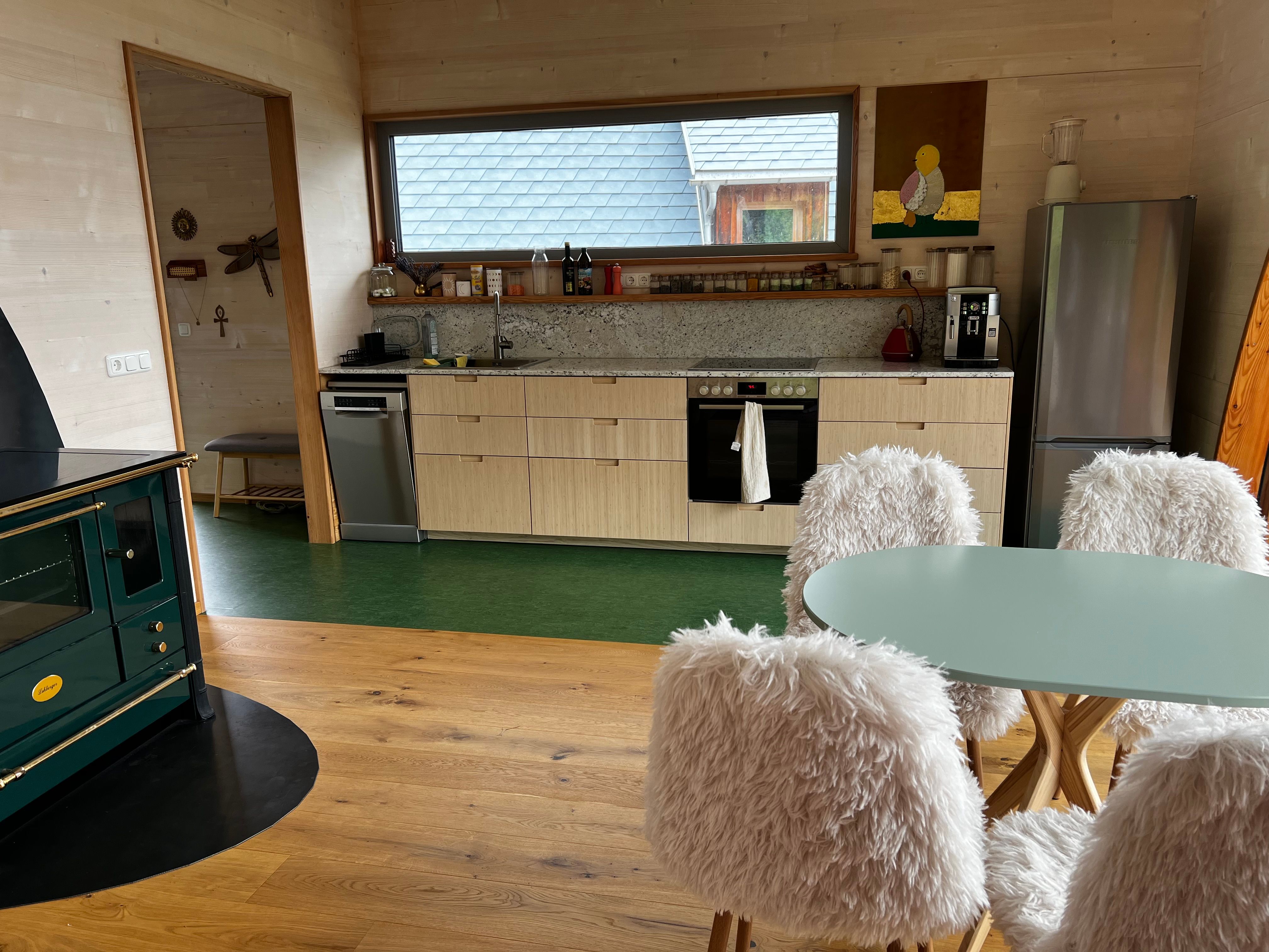 Moderne Küche mit Holzverkleidung, grüner Herd, Esstisch mit flauschigen Stühlen und großen Fenster mit Blick nach draußen.