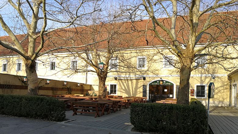 Ein gelbes Gebäude mit roten Dachziegeln und einem Biergarten mit Holzbänken und Tischen im Vordergrund.
