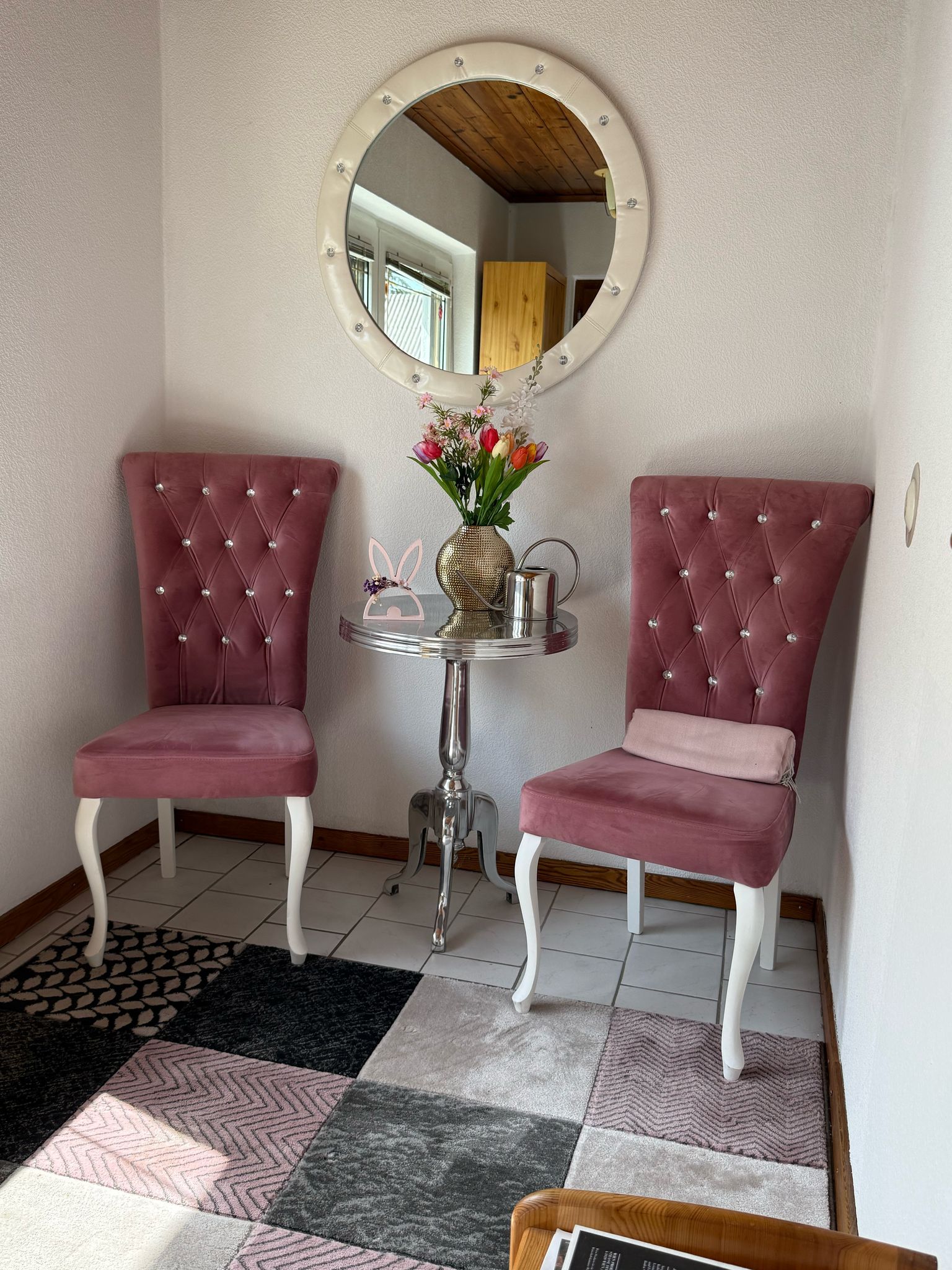 Ein Vorraum mit zwei rosa Stühlen, einem runden Spiegel und einem kleinen Tisch mit Blumen.