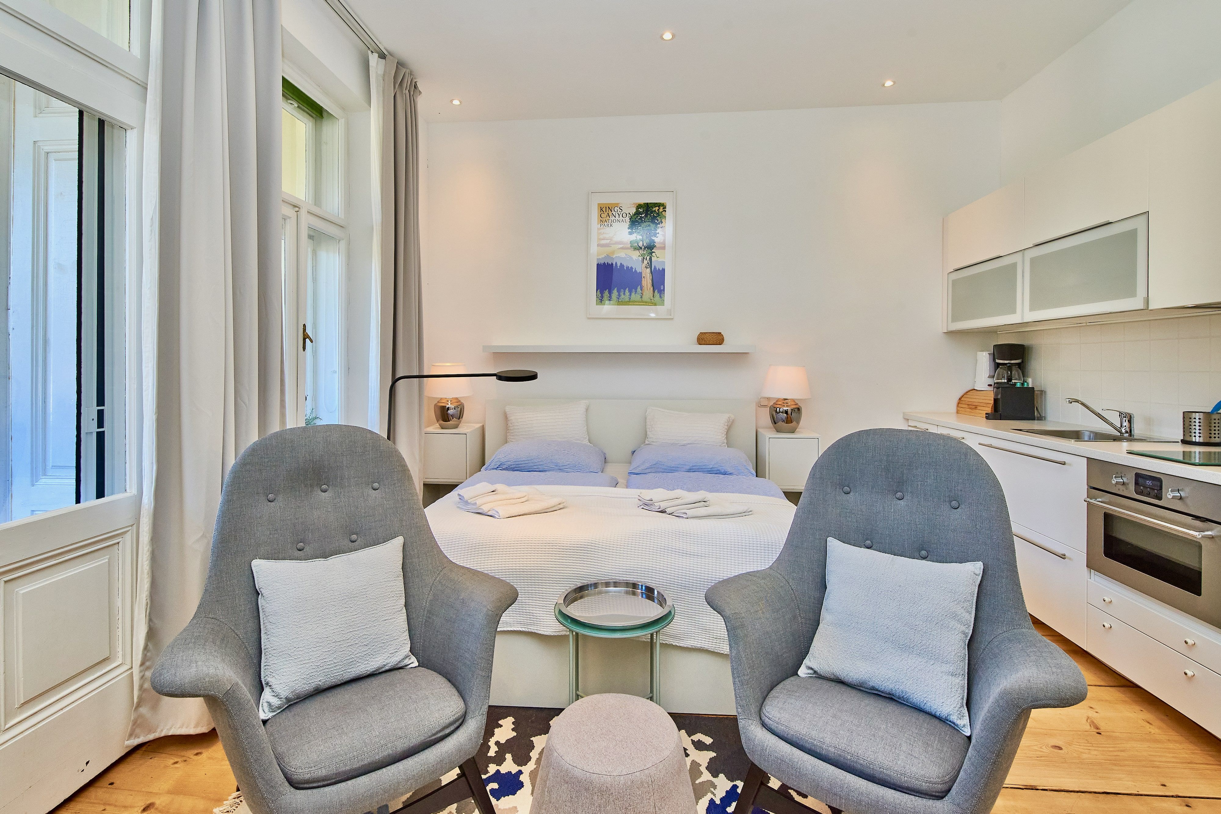 Modernes Studio-Apartment mit Doppelbett, zwei grauen Sesseln, Küchenzeile und großen Fenstern.