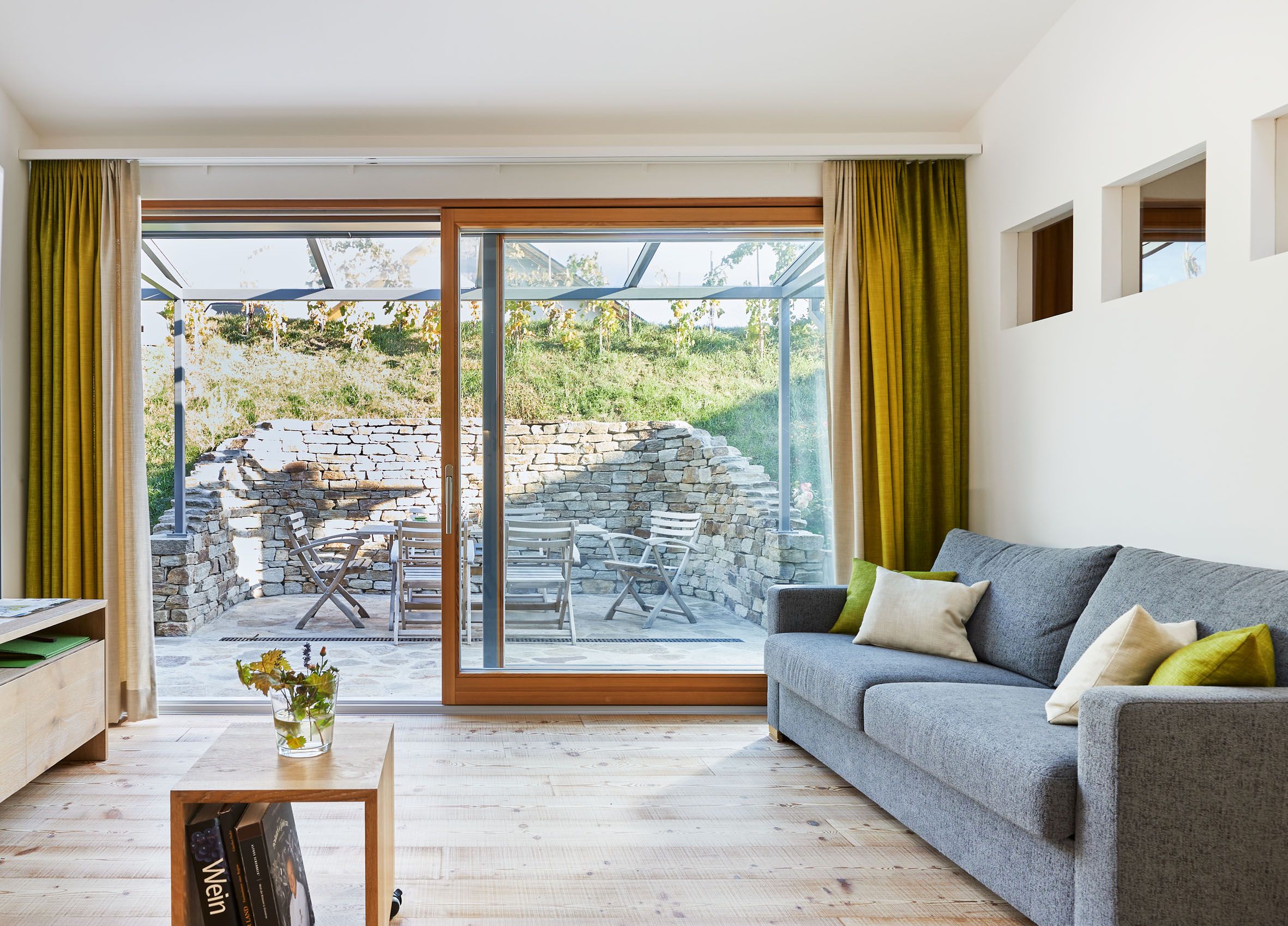 Modernes Wohnzimmer mit grauem Sofa, Holzboden und großen Fenstern mit Blick auf eine Terrasse mit Steinmauer und Gartenmöbeln.