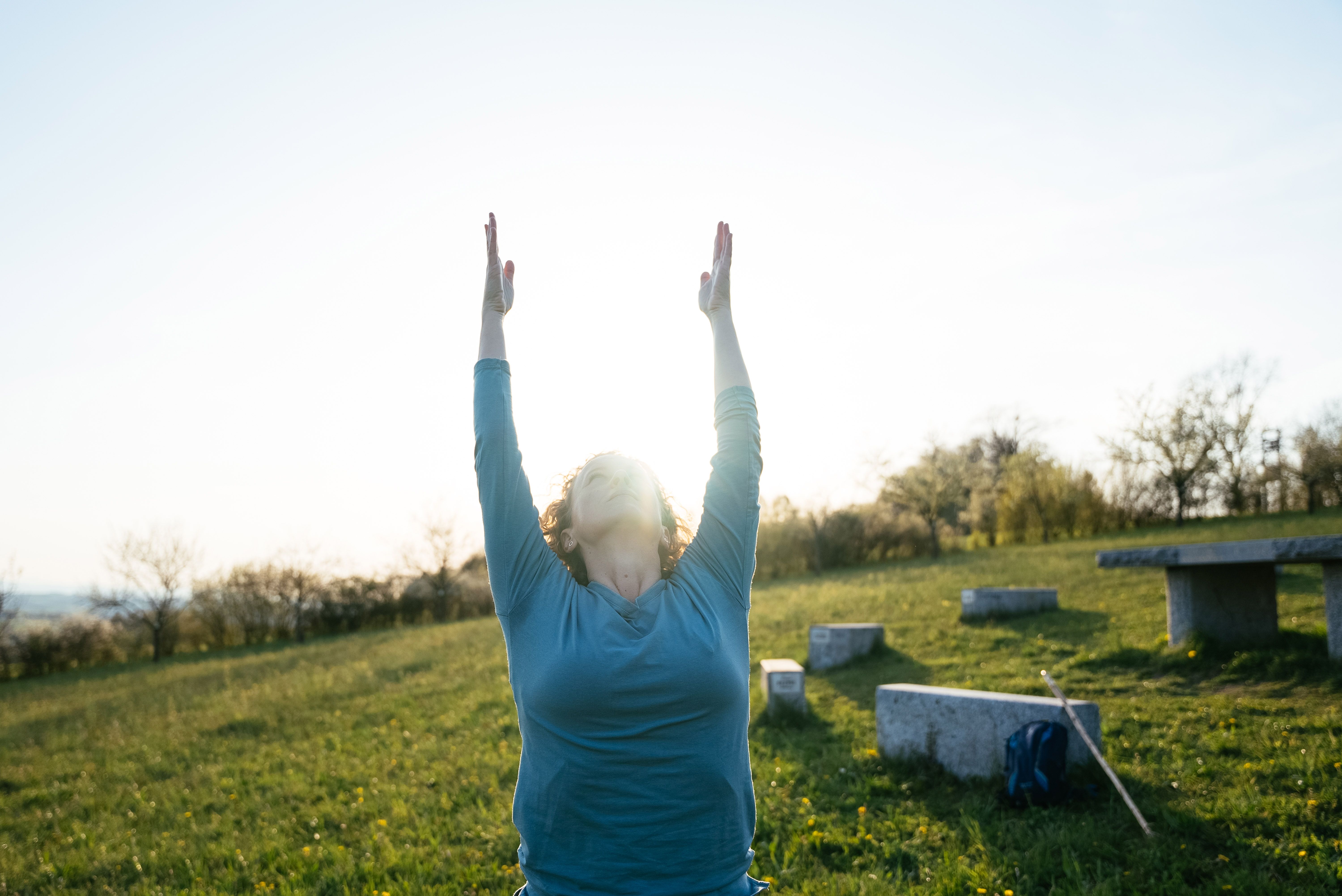 Wanderin streckt auf einer sonnigen Wiese veim Yoga beide Arme nach oben, während das Licht direkt hinter ihrem Gesicht strahlt.