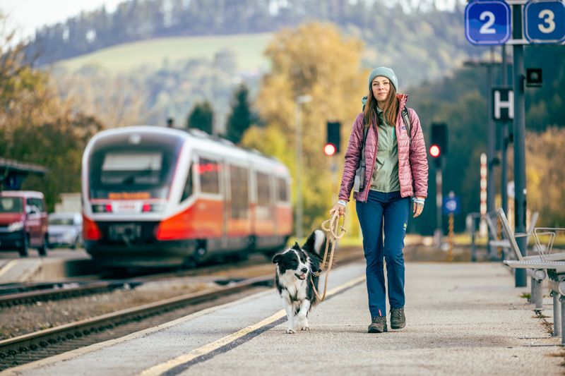 Frau mit Hund am Bahnhof Aspang, Zug im Hintergrund.