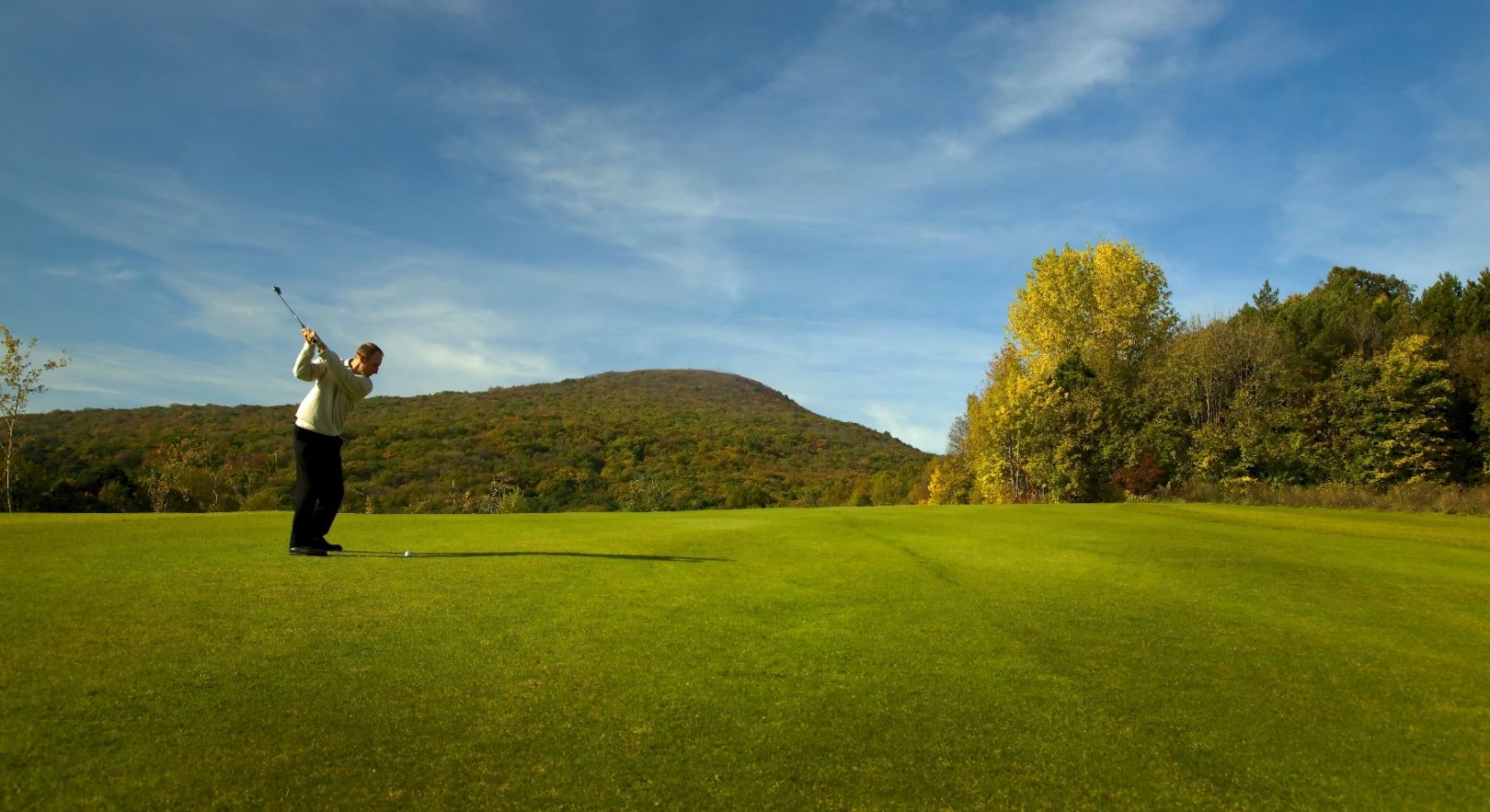 Golfer schlägt Ball auf einem grünen Golfplatz mit bewaldeten Hügeln im Hintergrund.
