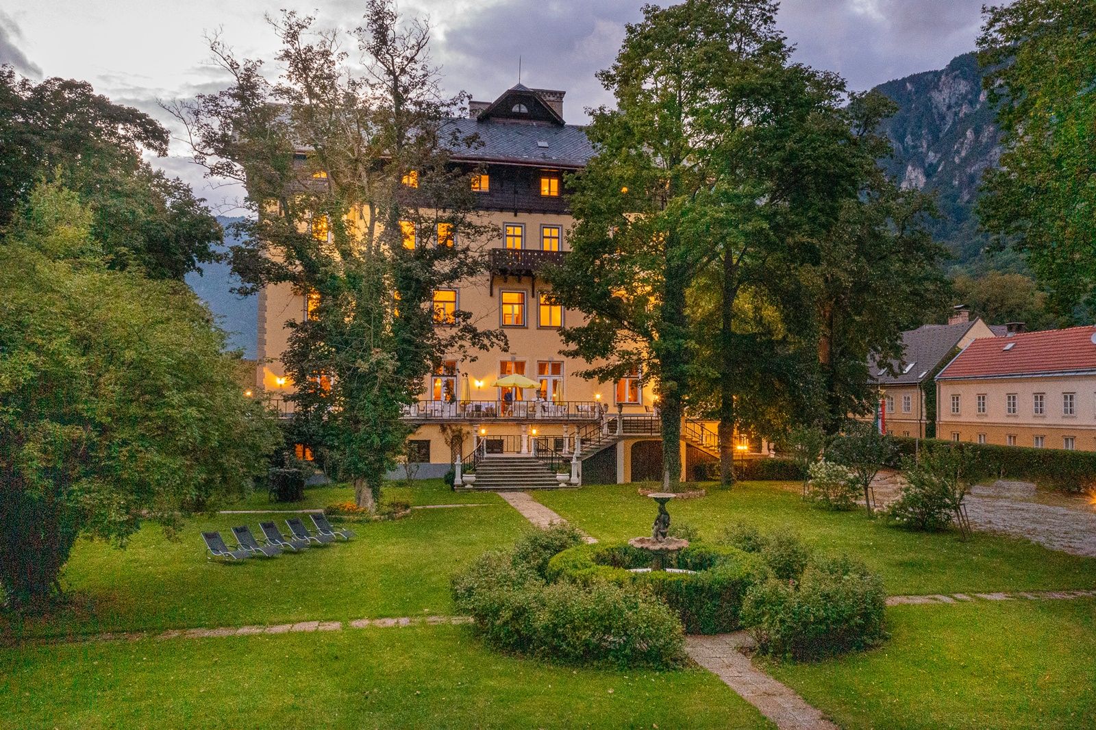 Ein beleuchtetes Gebäude mit Garten und Liegestühlen bei Dämmerung.