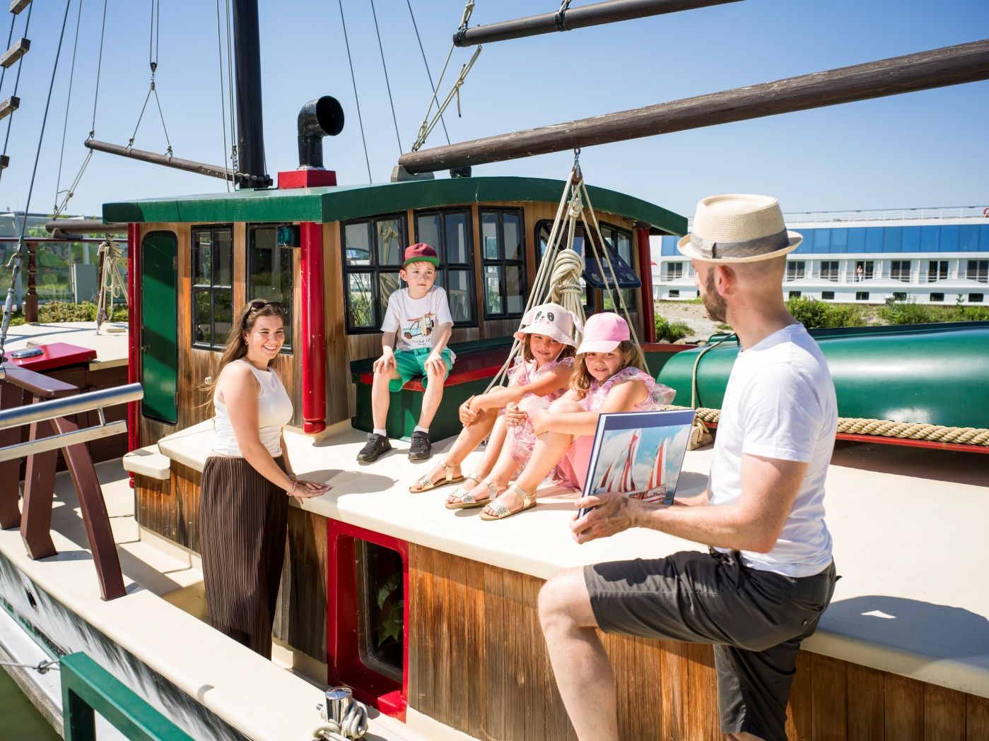 Kinder sitzen am Deck des historischem Hundertwasser-Schiffs, Frau steht daneben und der Führer zeigt Fotos des Schiffes