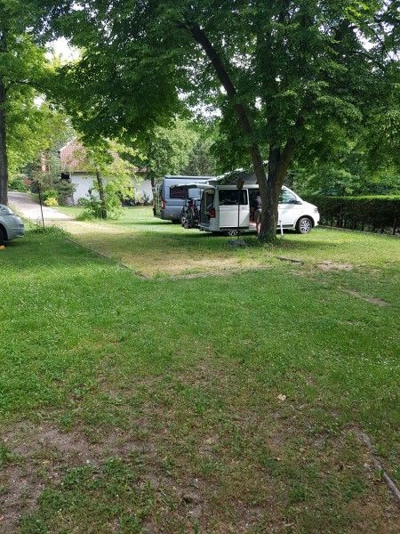 Zwei Wohnmobile stehen auf einem grünen Campingplatz unter Bäumen.