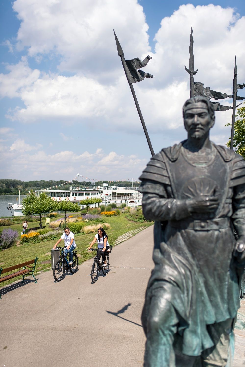Zwei Radfahrer fahren auf einem Weg entlang der Donau in Tulln. Im Vordergrund steht eine Statue, im Hintergrund ist ein Flusskreuzfahrtschiff zu sehen.
