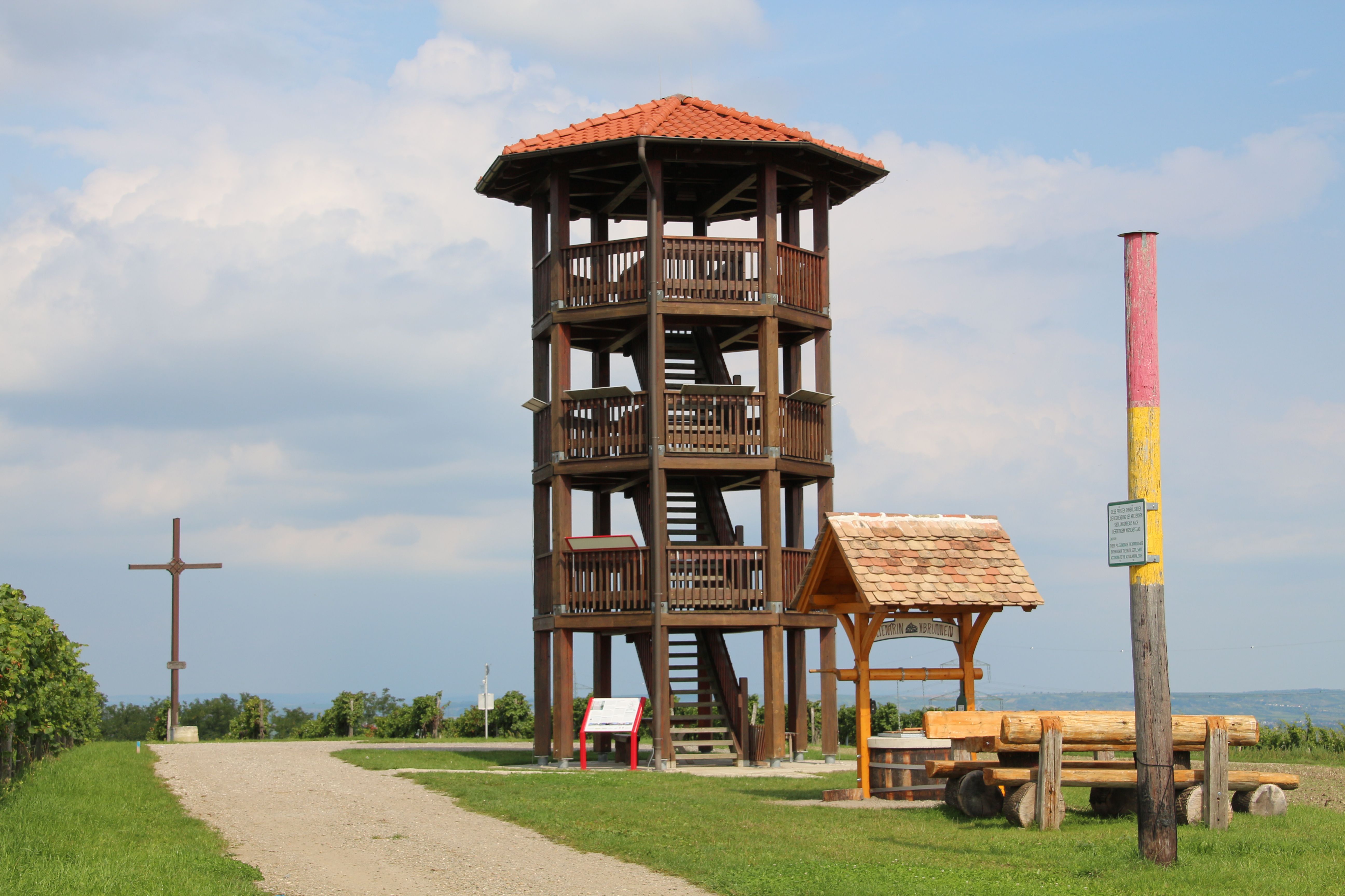 Holzaussichtsturm mit rotem Dach auf einer Wiese, daneben ein Holzkreuz und eine Sitzbank.