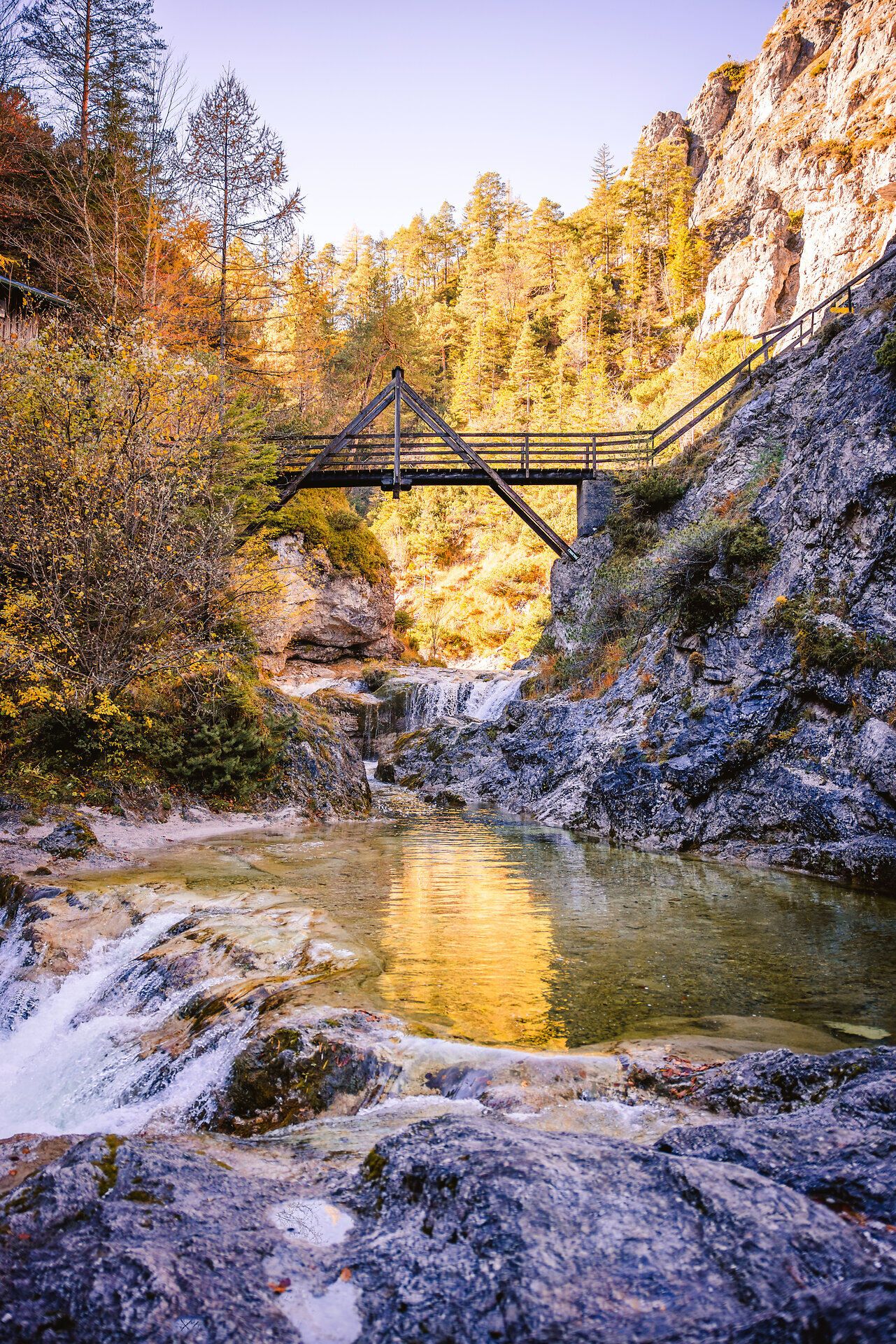 Die goldenen Herbstfarben spiegeln sich im klaren Wasser des Baches wider, während die sanften Geräusche des plätschernden Wassers eine friedliche Atmosphäre schaffen. Eine malerische Holzbrücke verbindet die beiden Ufer und lädt dazu ein, die atemberaubende Natur des Naturparks Ötscher Tormäuer zu erkunden.