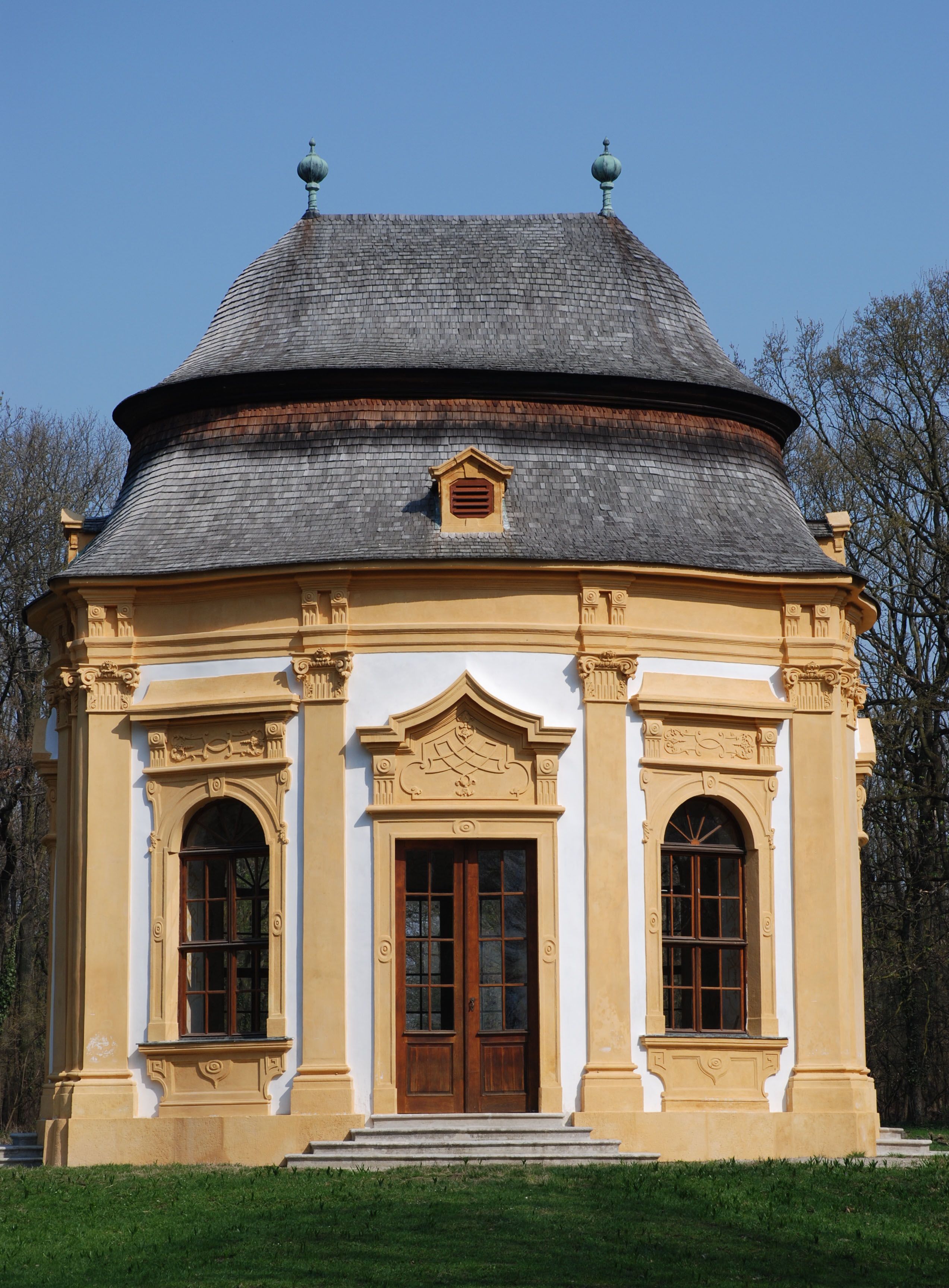 Barocker Gartenpavillon mit gelben Verzierungen und grauem Dach vor blauem Himmel.