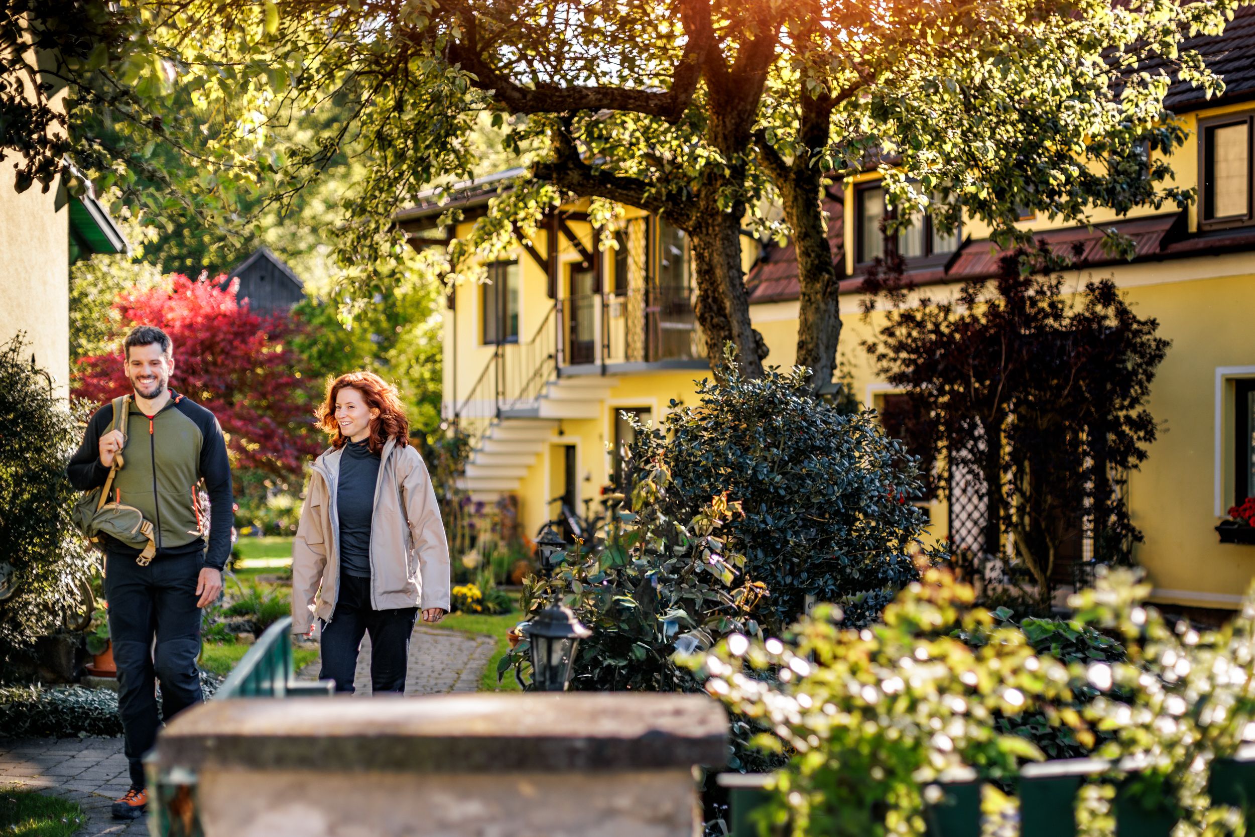 Ein Mann und eine Frau in Wanderkleidung gehen durch den grünen, gepflegten Garten der Pension Hendling
