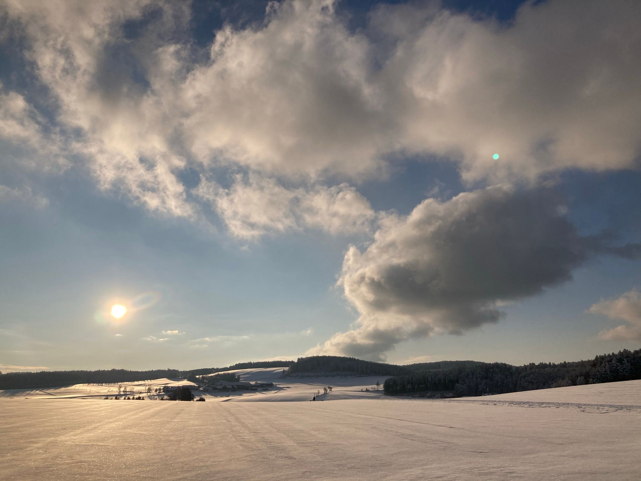 Verschneite Landschaft unter bewölktem Himmel mit tief stehender Sonne.