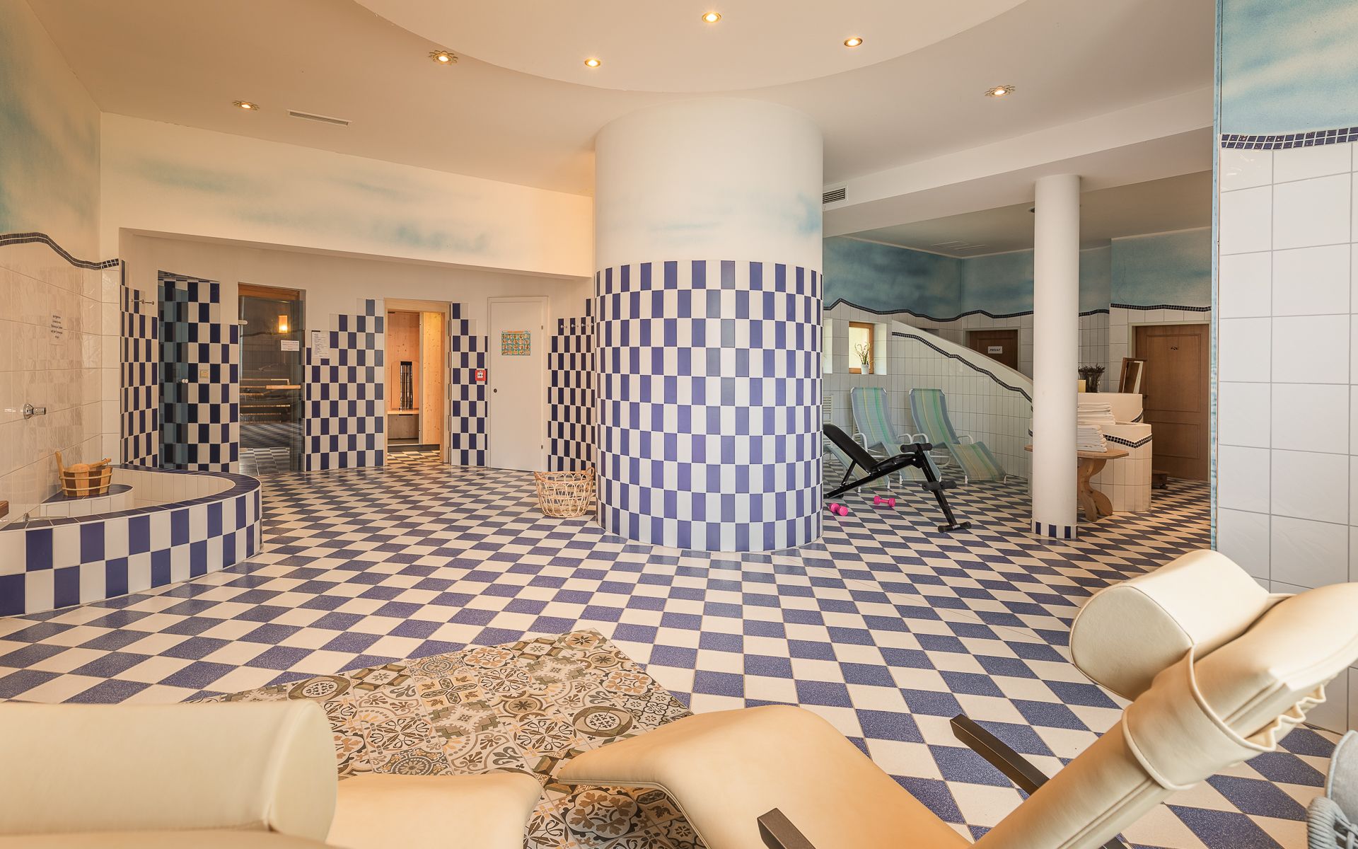 Innenansicht eines Saunabereichs mit blau-weiß karierten Fliesen, Liegestühlen und Fitnessgeräten.
