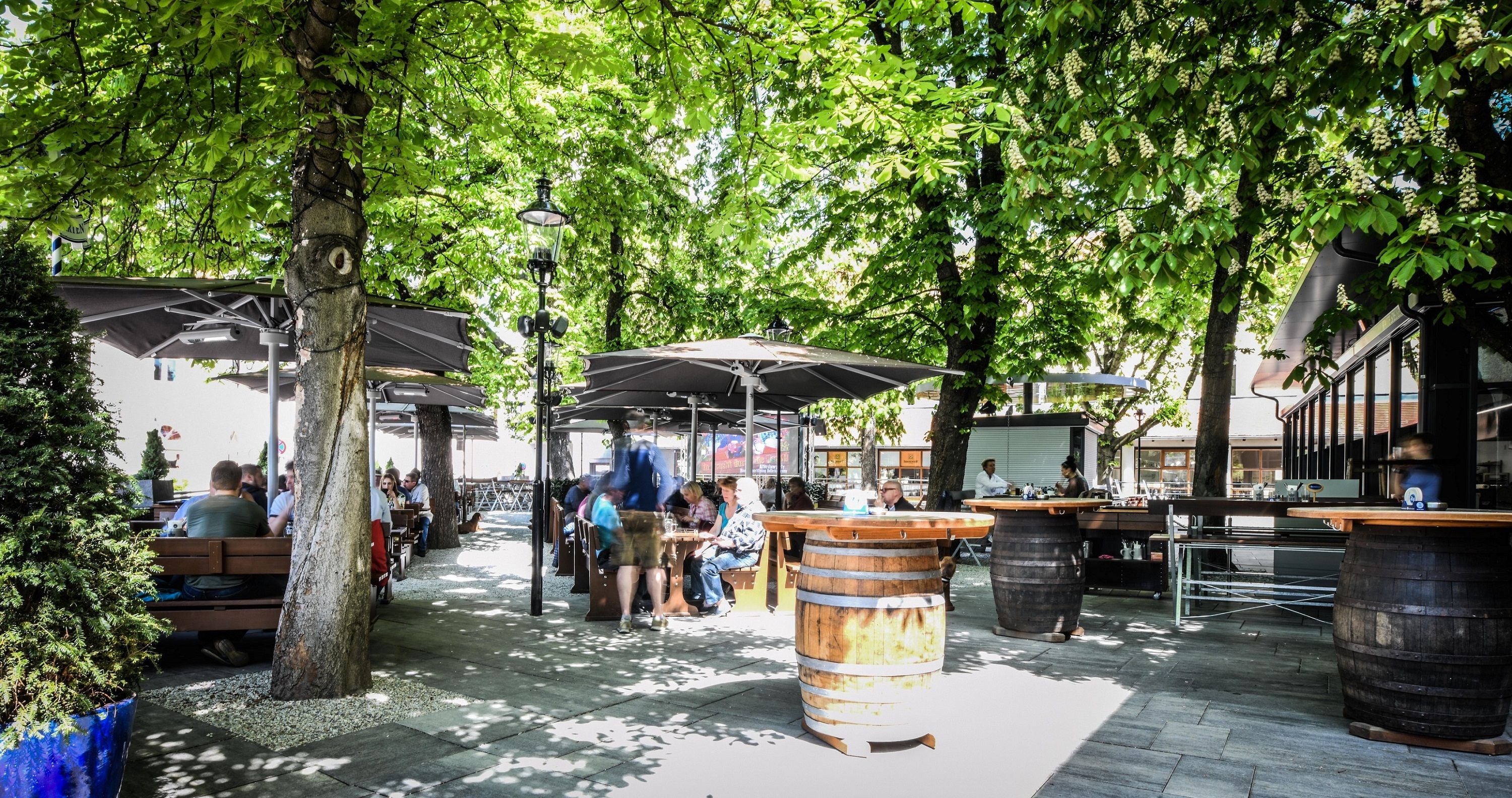 Ein Biergarten mit Holztischen und -fässern unter grünen Bäumen, Menschen sitzen und genießen die Atmosphäre.