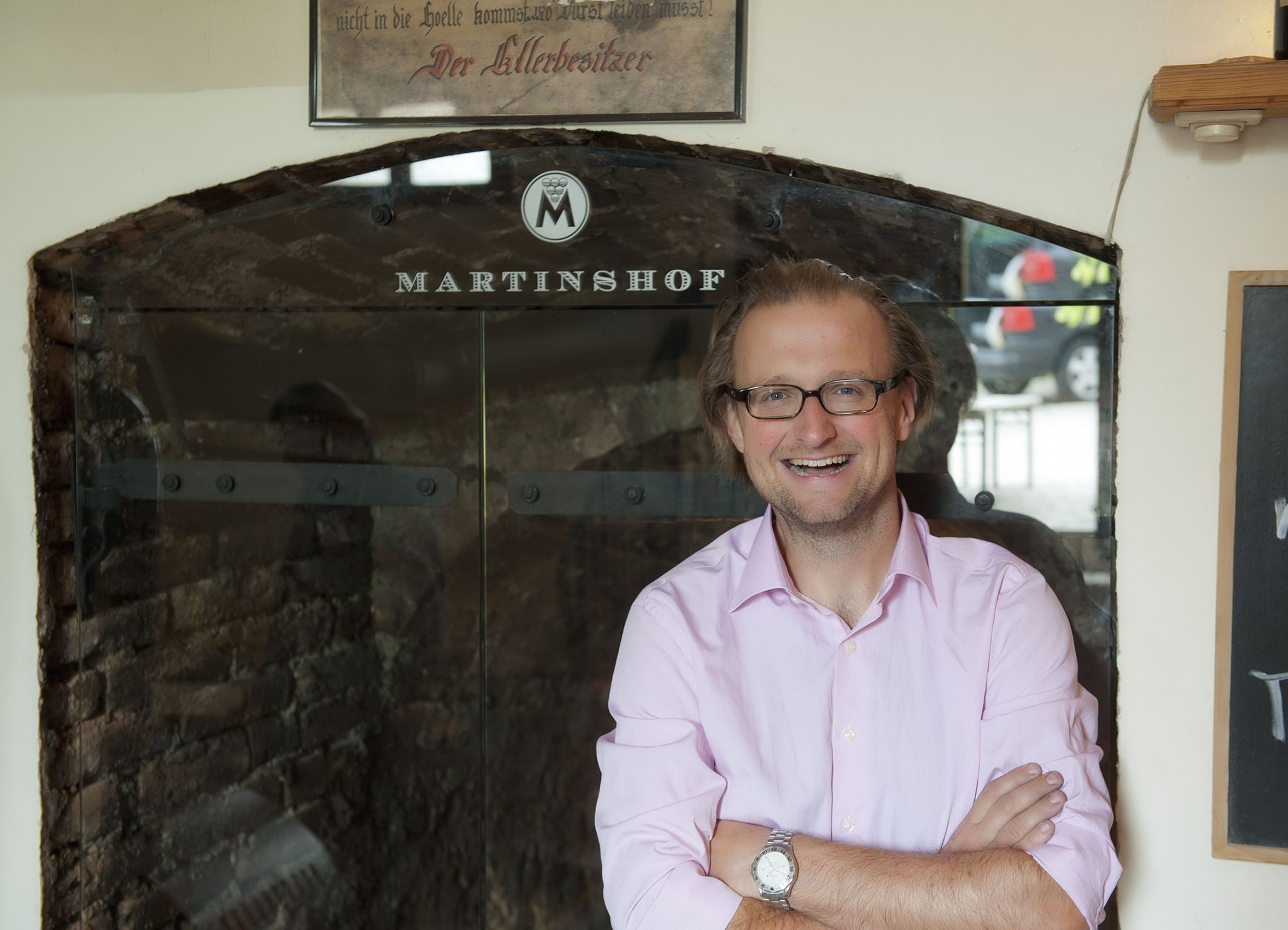 Ein Mann mit Brille und rosa Hemd steht lächelnd vor einem Schild mit der Aufschrift 'Martinshof'.