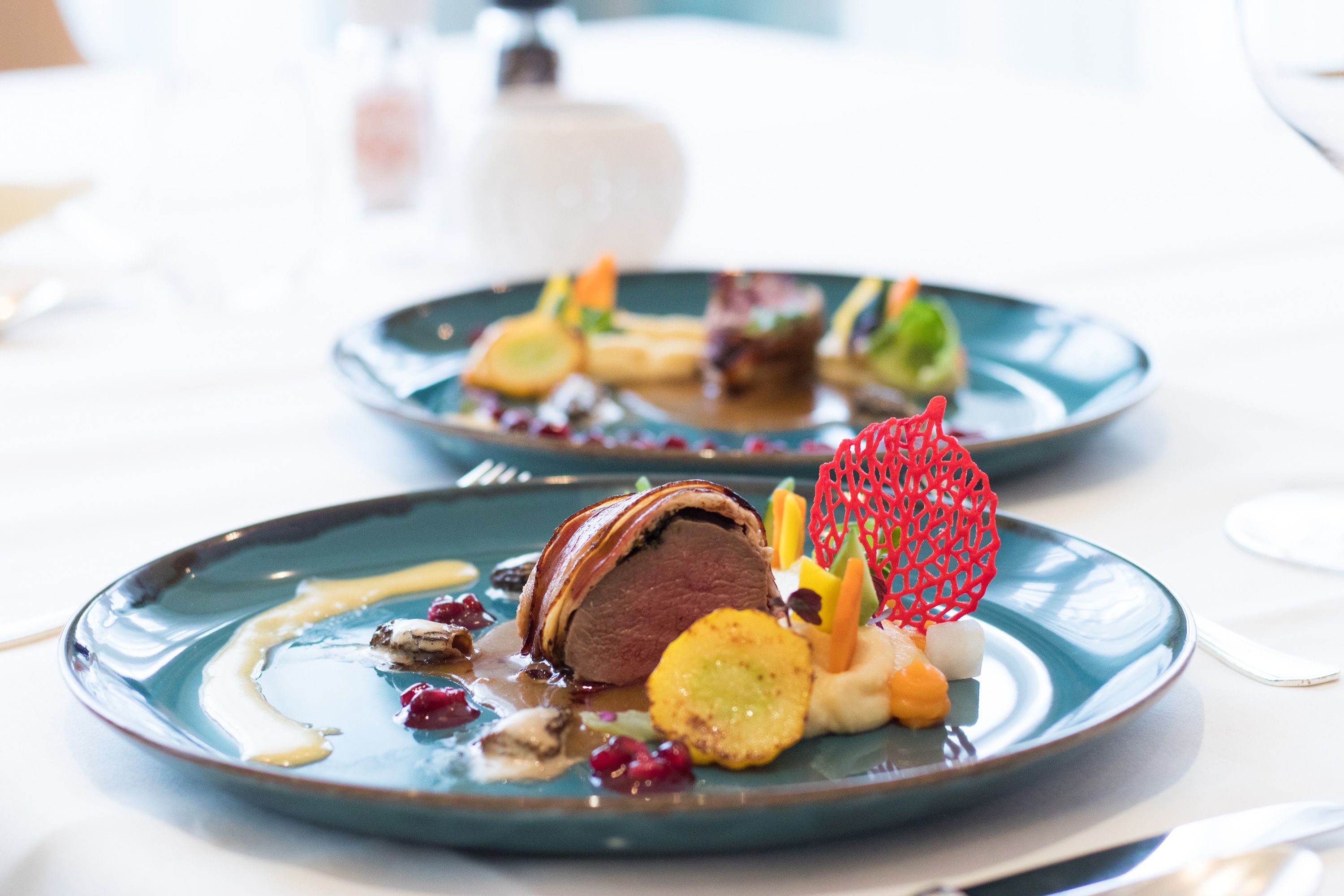 Gourmetgericht auf einem blauen Teller, kunstvoll angerichtet mit Fleisch, Gemüse und einer roten Dekoration.