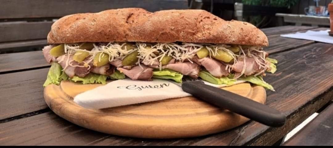 Ein großes Sandwich mit Fleisch, Salat, Gurken und Käse auf einem Holzbrett, daneben ein Messer mit Serviette.