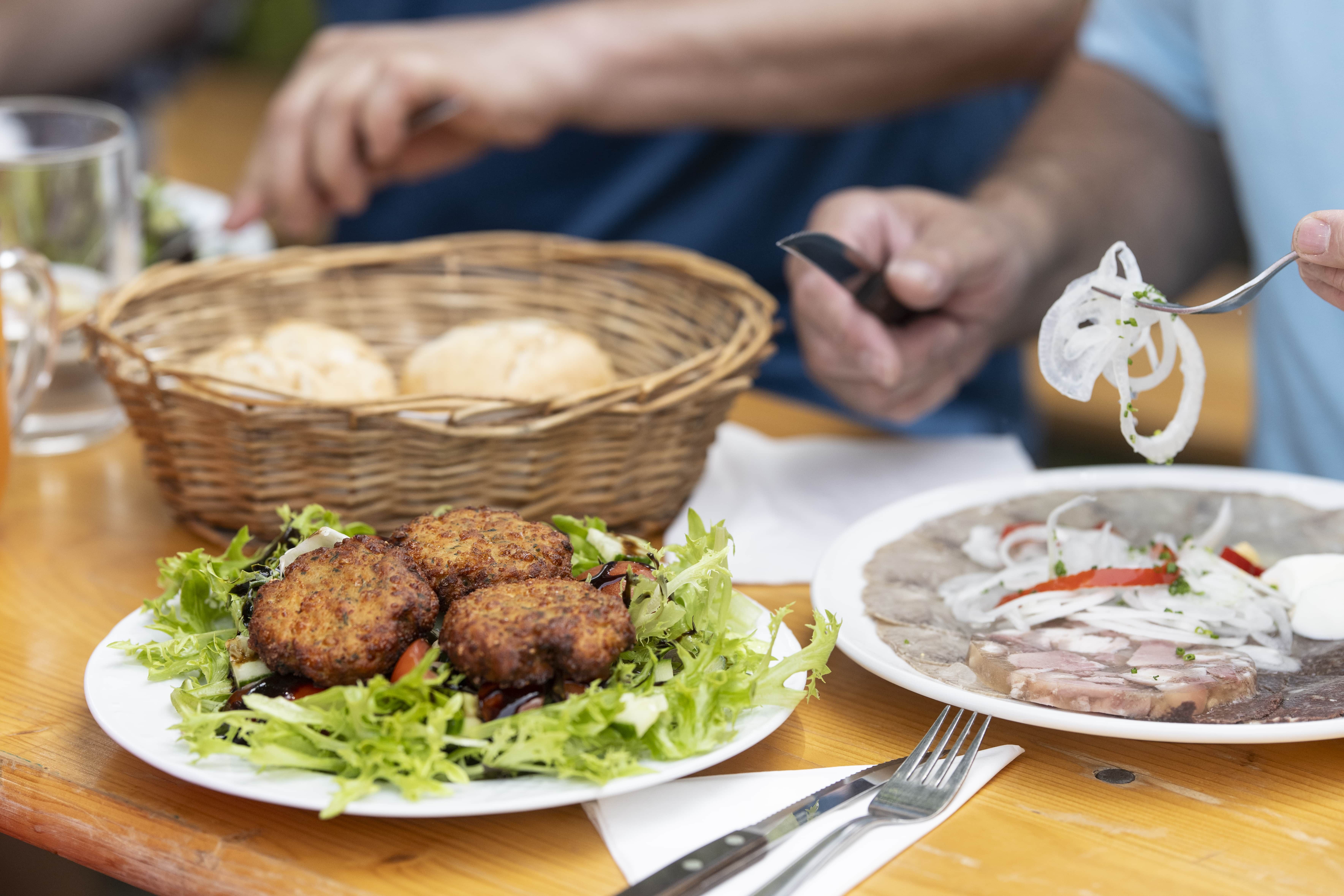 Ein Tisch mit einem Teller voller Fleischlaberl auf Salat, einem Korb mit Gebäck und einem Teller mit Presswurst und Zwiebeln.