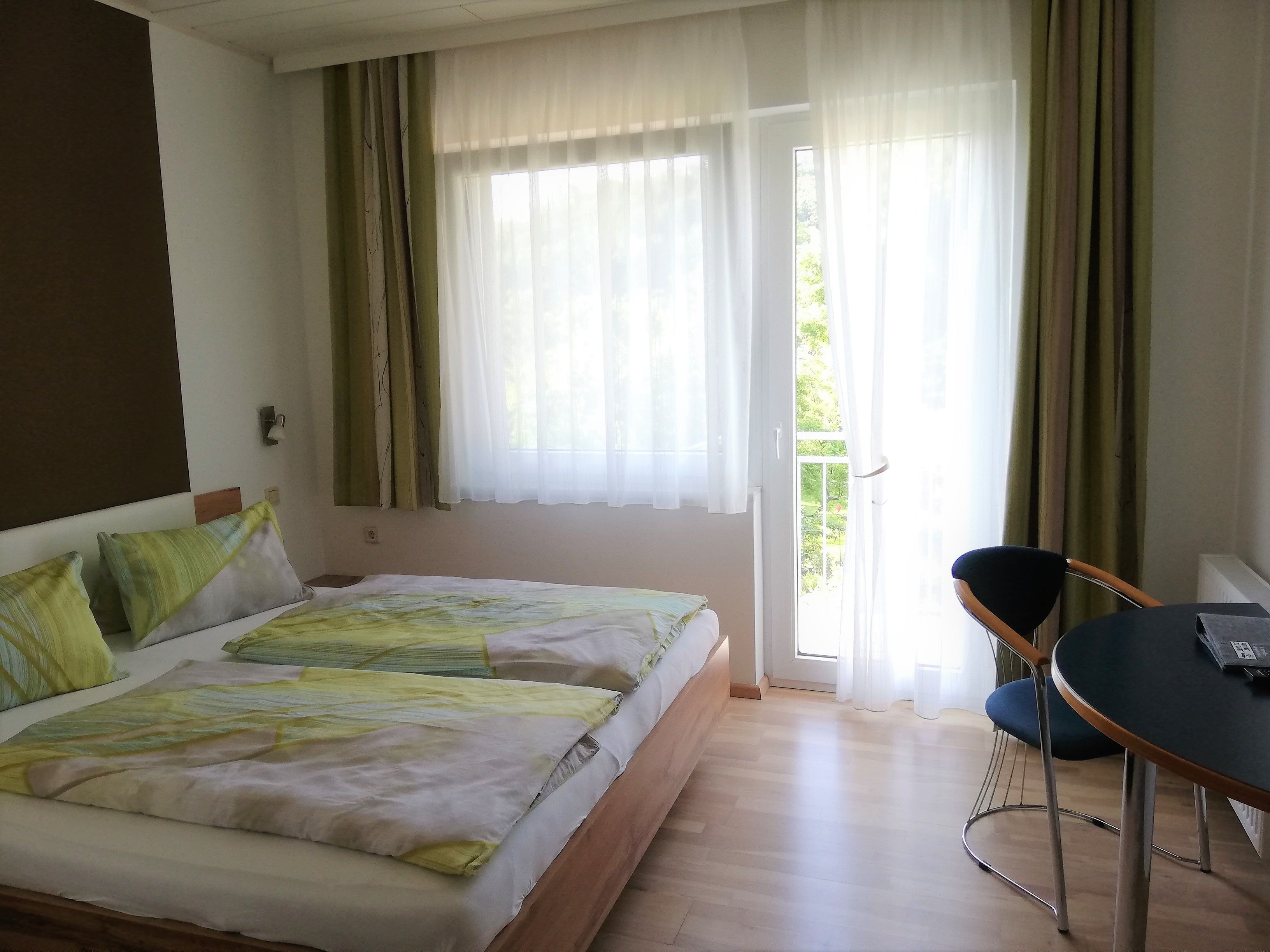 Ein Doppelzimmer mit einem Bett, einem Tisch und einem Stuhl. Ein Balkon ist durch eine Glastür sichtbar.