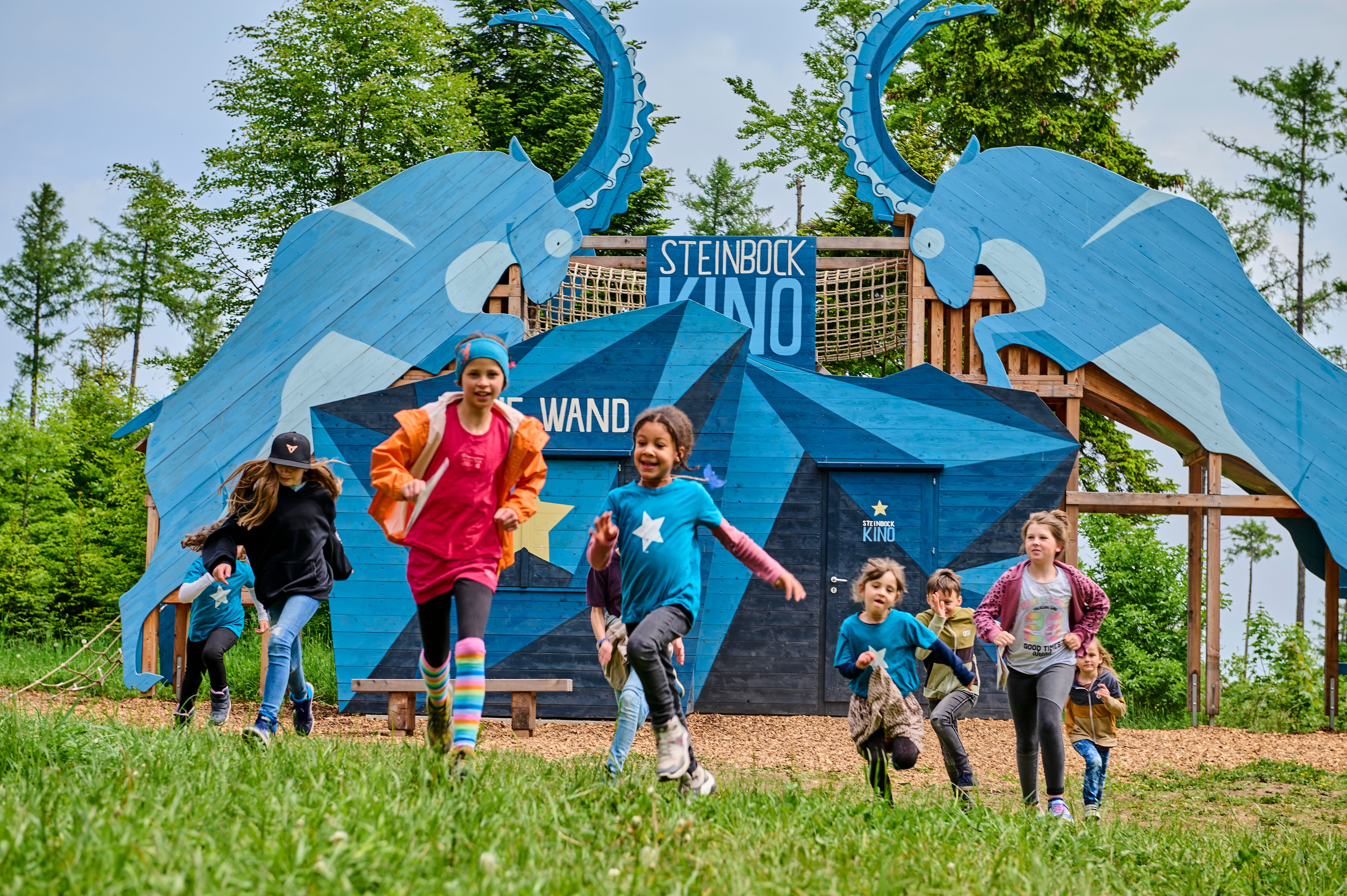 Kinder rennen vor einer blauen Holzkonstruktion mit Steinbock-Motiven und der Aufschrift 'Steinbock Kino'.