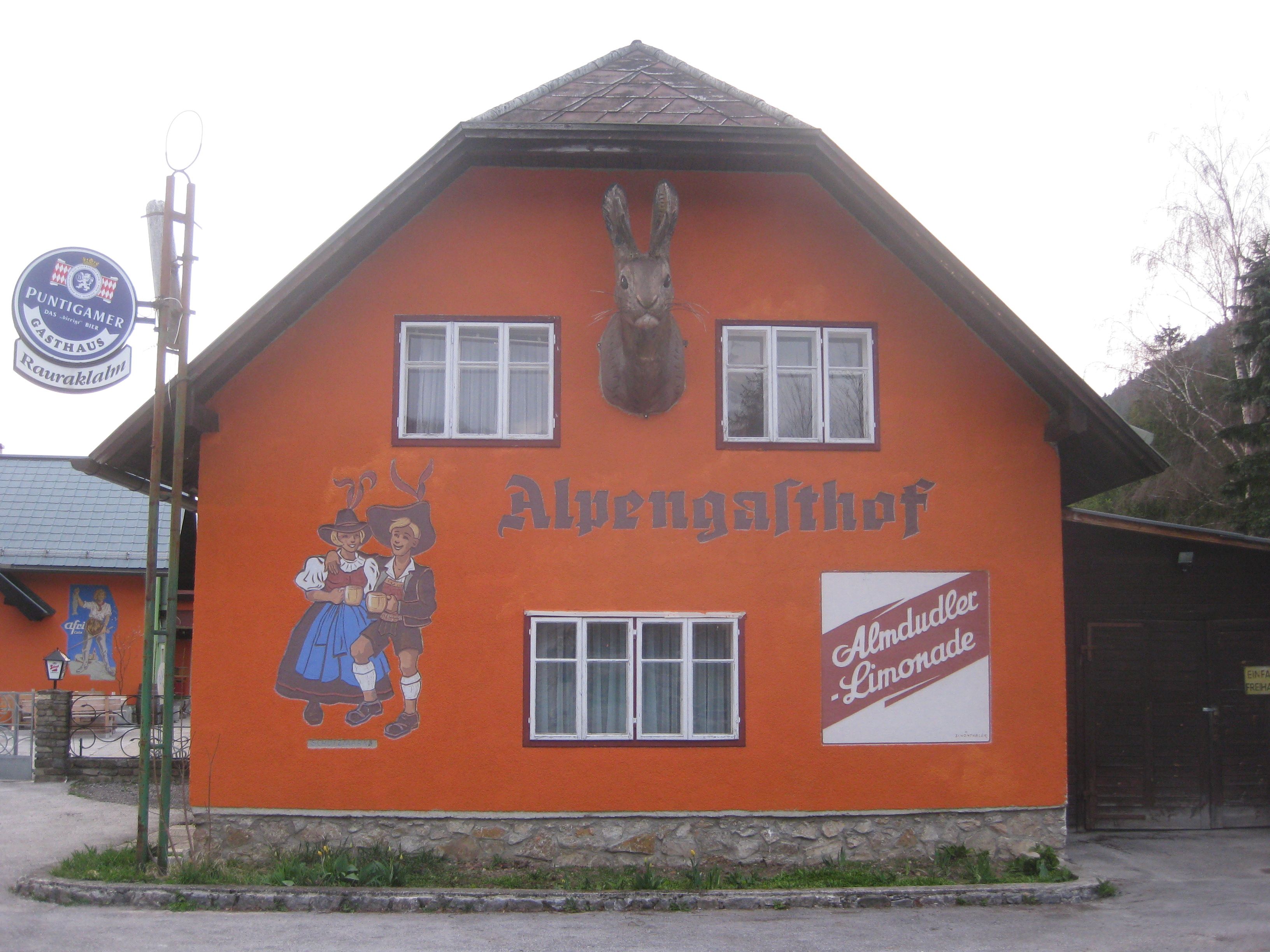 Orange gestrichenes Gebäude mit der Aufschrift 'Alpengasthof', Wandmalerei von tanzendem Paar und Almdudler-Werbung.