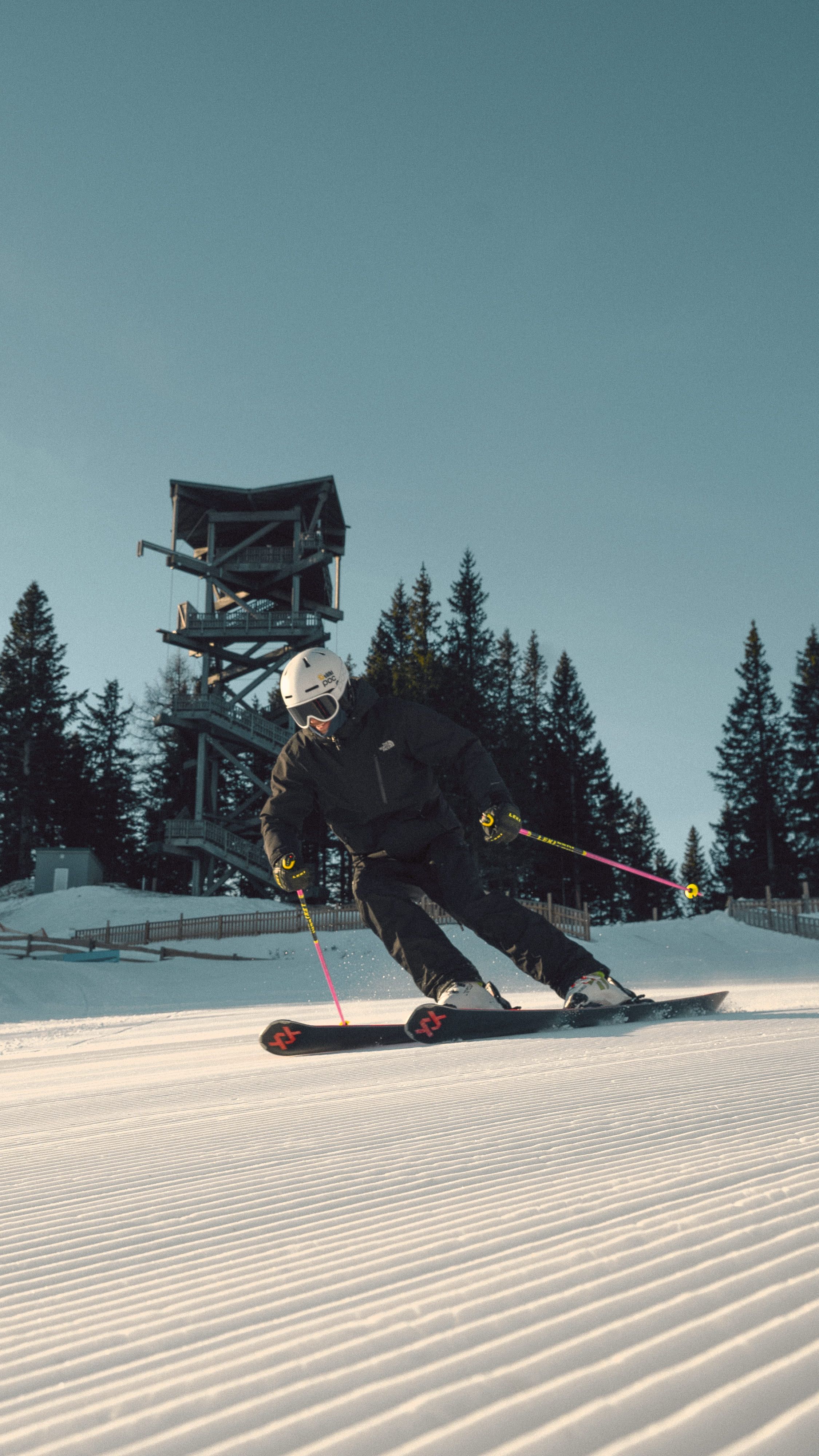 Skifahrer in schwarzer Kleidung fährt auf präparierter Piste am Semmering Hirschenkogel, im Hintergrund ein Aussichtsturm und Bäume.