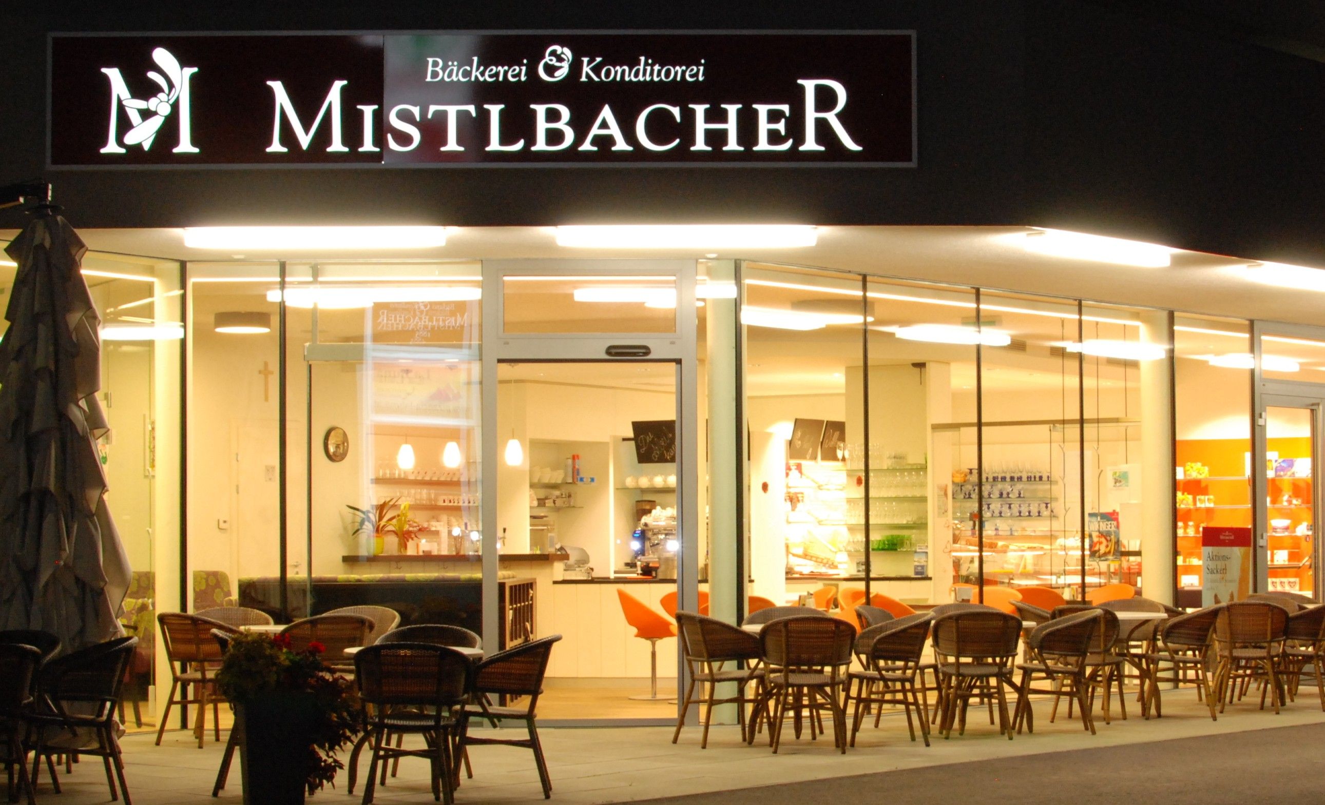 Außenansicht eines Cafés mit dem Namen Mistelbacher, beleuchtet und mit Sitzgelegenheiten im Freien.