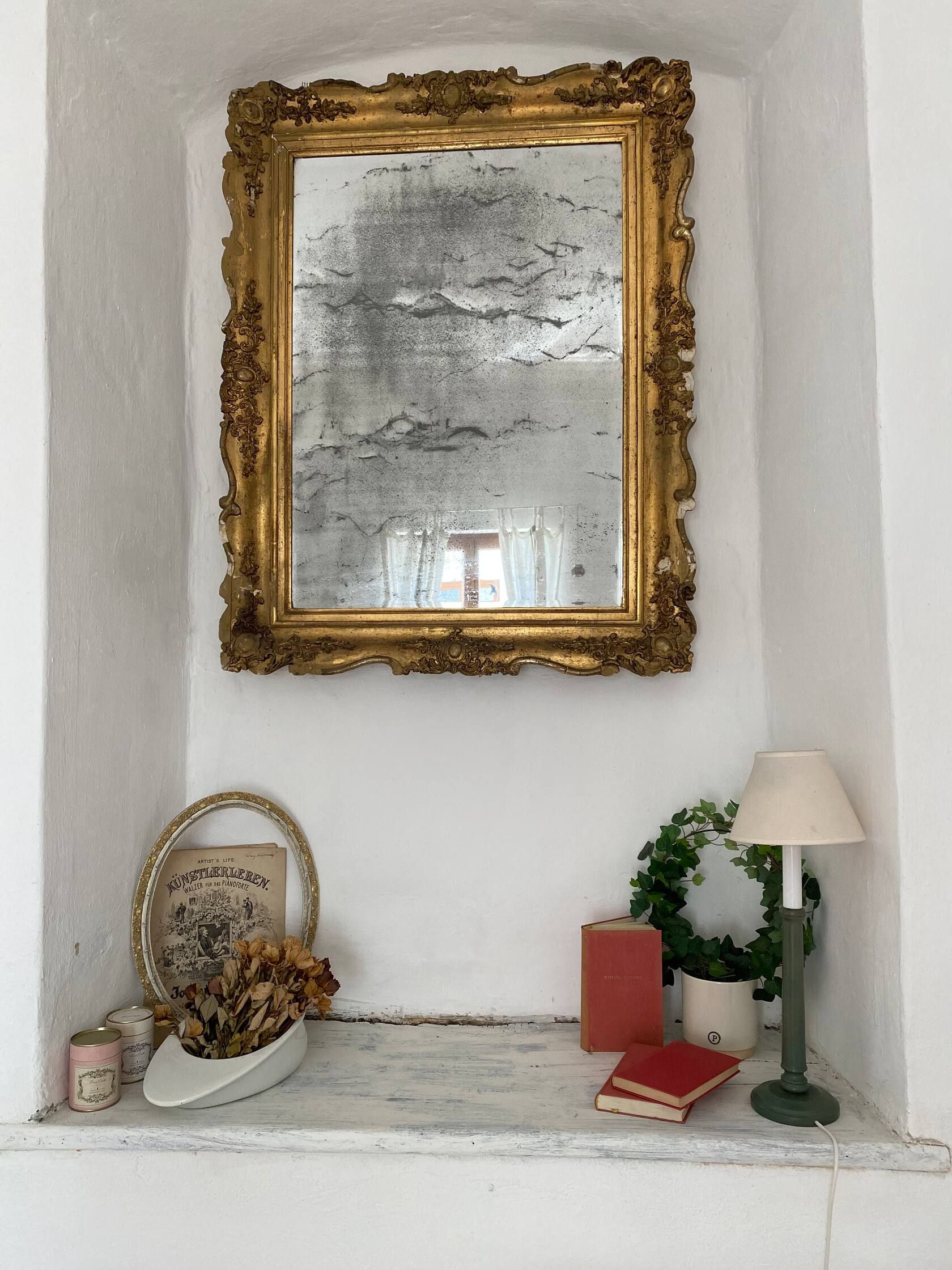 Ein antiker Spiegel mit goldener Verzierung über einer Fensterbank mit Deko.