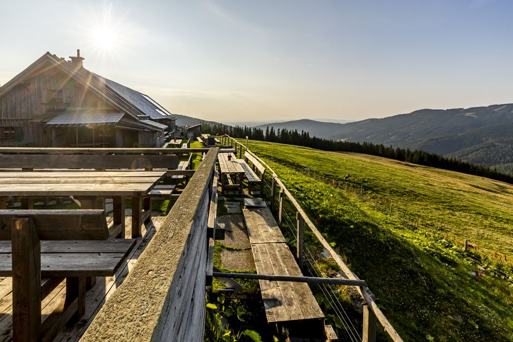 Eine Berghütte mit Holzbänken und Tischen, umgeben von grünen Wiesen und Bergen im Sonnenlicht.
